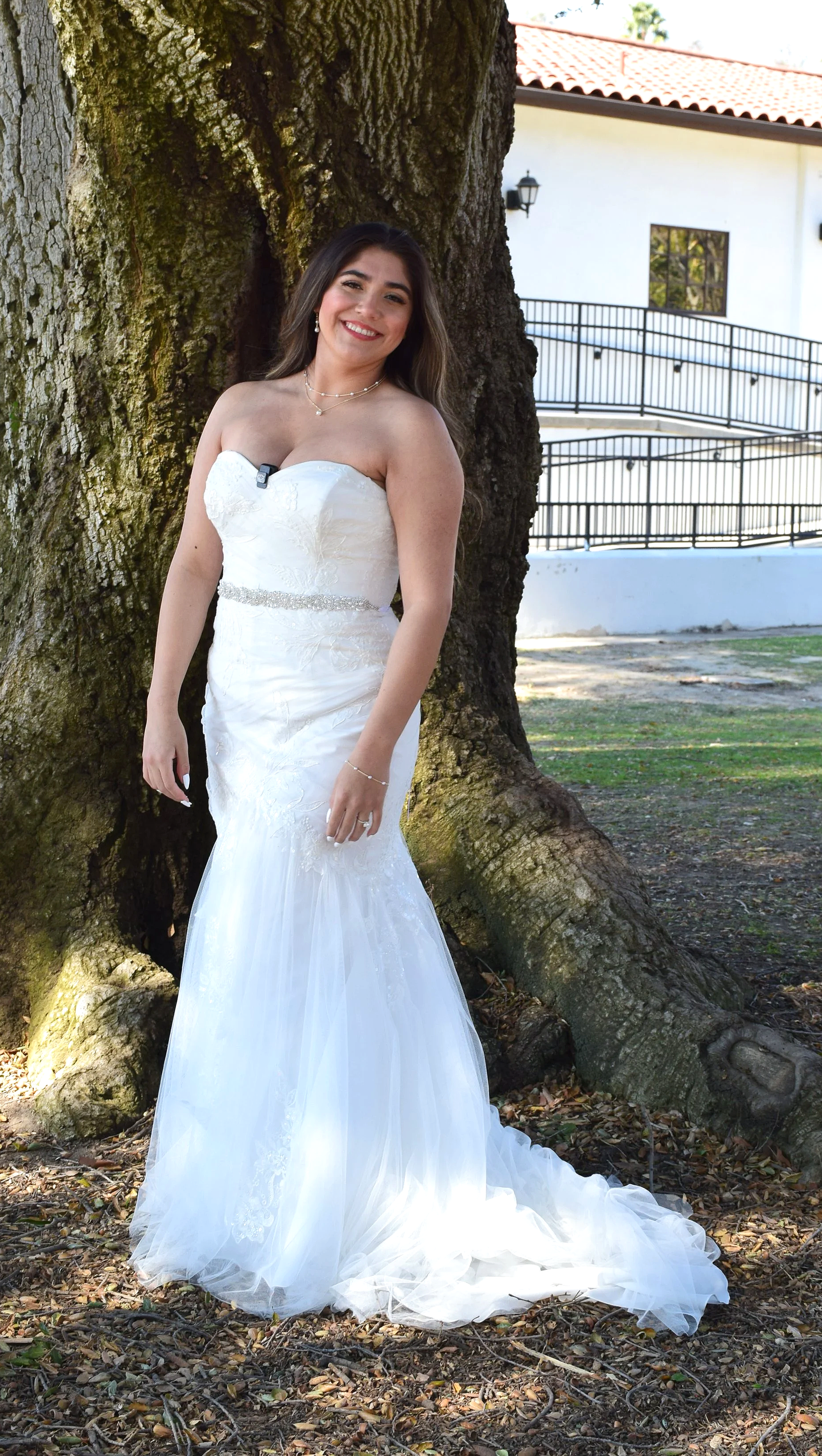 A smiling woman in a white wedding dress standing outdoors in front of a large tree with textured bark. She is wearing jewelry and leaning slightly against the tree.