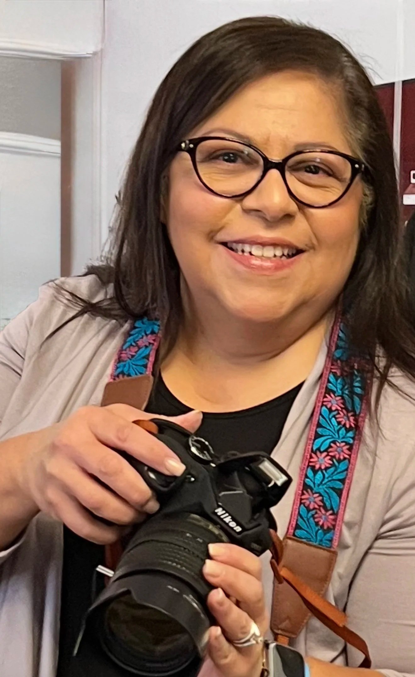 A smiling woman with dark brown hair, wearing glasses, holding a professional camera, and wearing a colorful strap around her neck, standing indoors.
