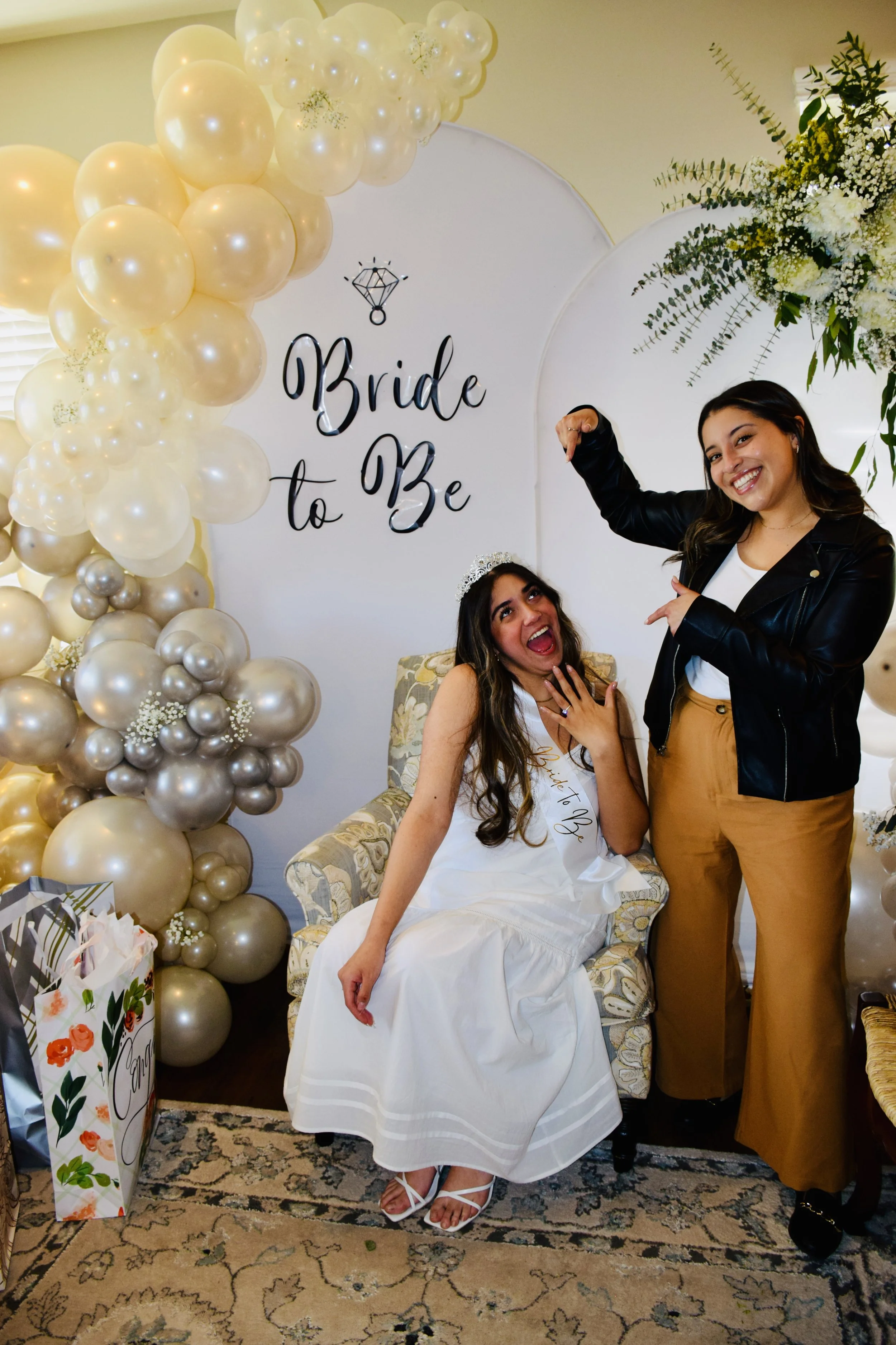 A woman in a white dress sitting on a floral armchair, with a sash reading 'Bride to Be', celebrating her bridal shower, next to a woman in a black leather jacket and tan pants, both smiling and pointing at each other. The background features a white circular sign with 'Bride to Be' written on it, decorated with balloons in shades of cream, white, and champagne, and a floral arrangement of white flowers and greenery.