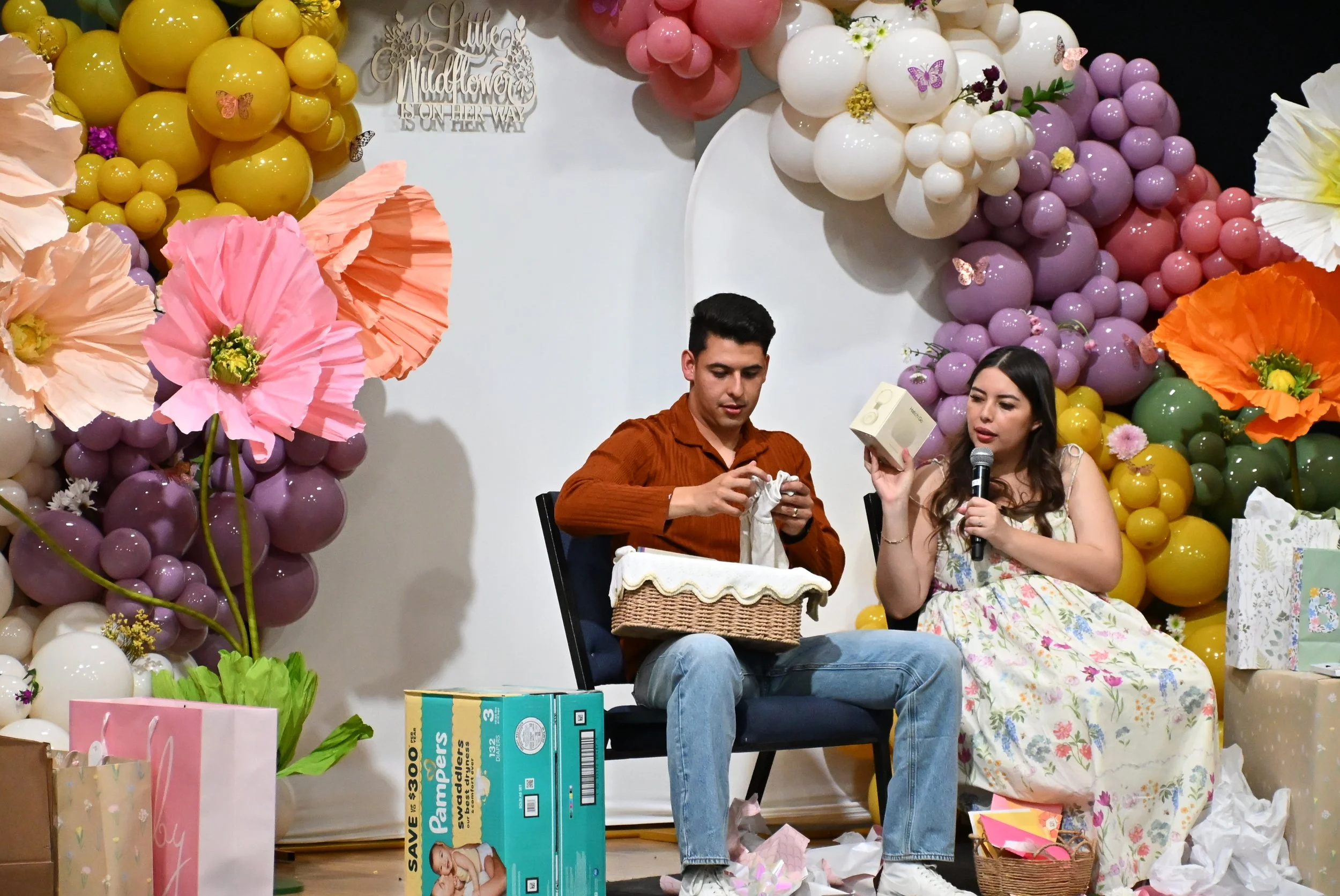 A decorated celebration scene with balloons in yellow, pink, purple, white, and orange, large paper flowers, and gifts. Two people, a man and a woman, are seated on stage, with the woman holding a microphone and the man holding gift wrapping material