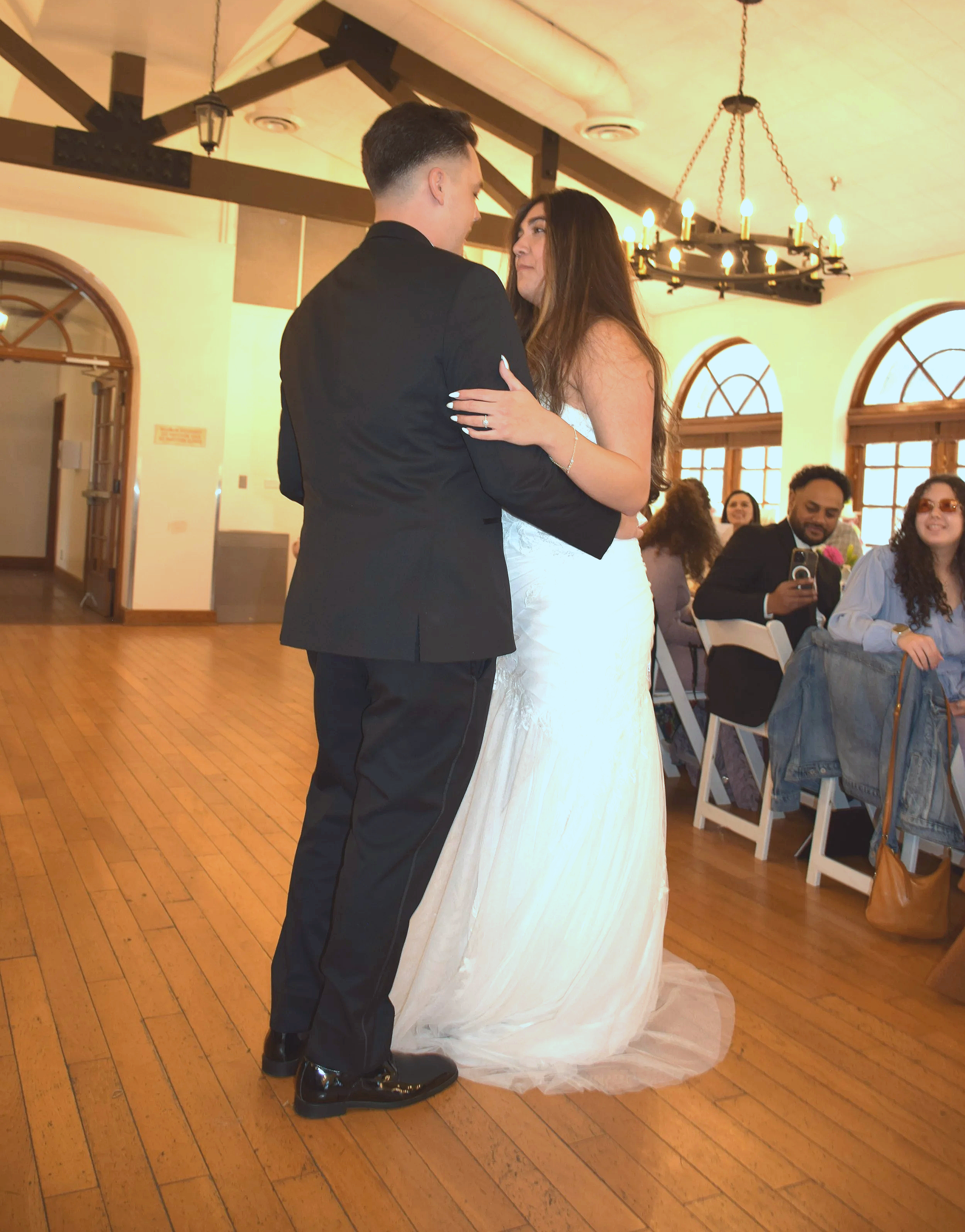 A bride and groom are dancing during their wedding reception, with guests seated at tables in the background.