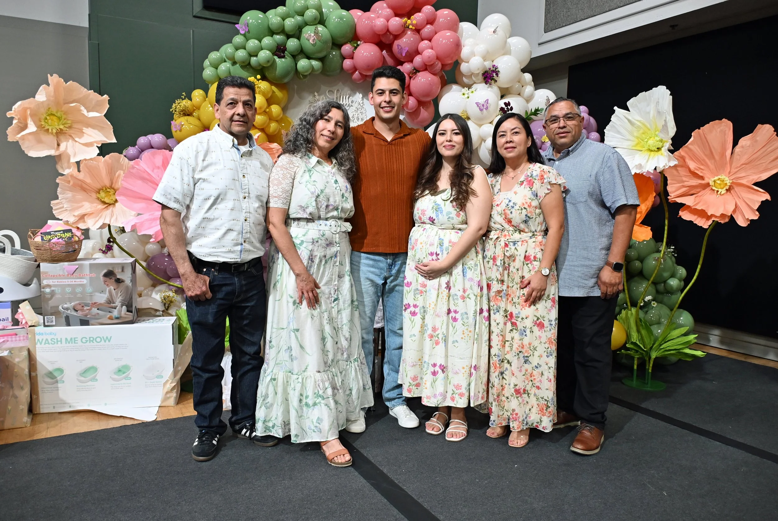 A group of six people standing together at a celebration event, with a colorful balloon and flower decoration backdrop.
