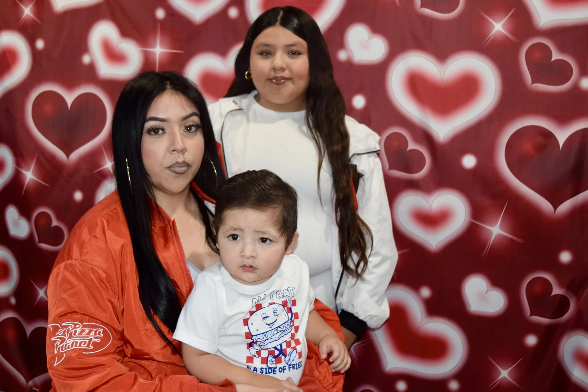A woman with long dark hair, wearing an orange jacket, sitting with two children in front of a backdrop with red and white heart shapes. One child is a girl with long dark hair, and the other is a boy with short dark hair and a white shirt with a ham