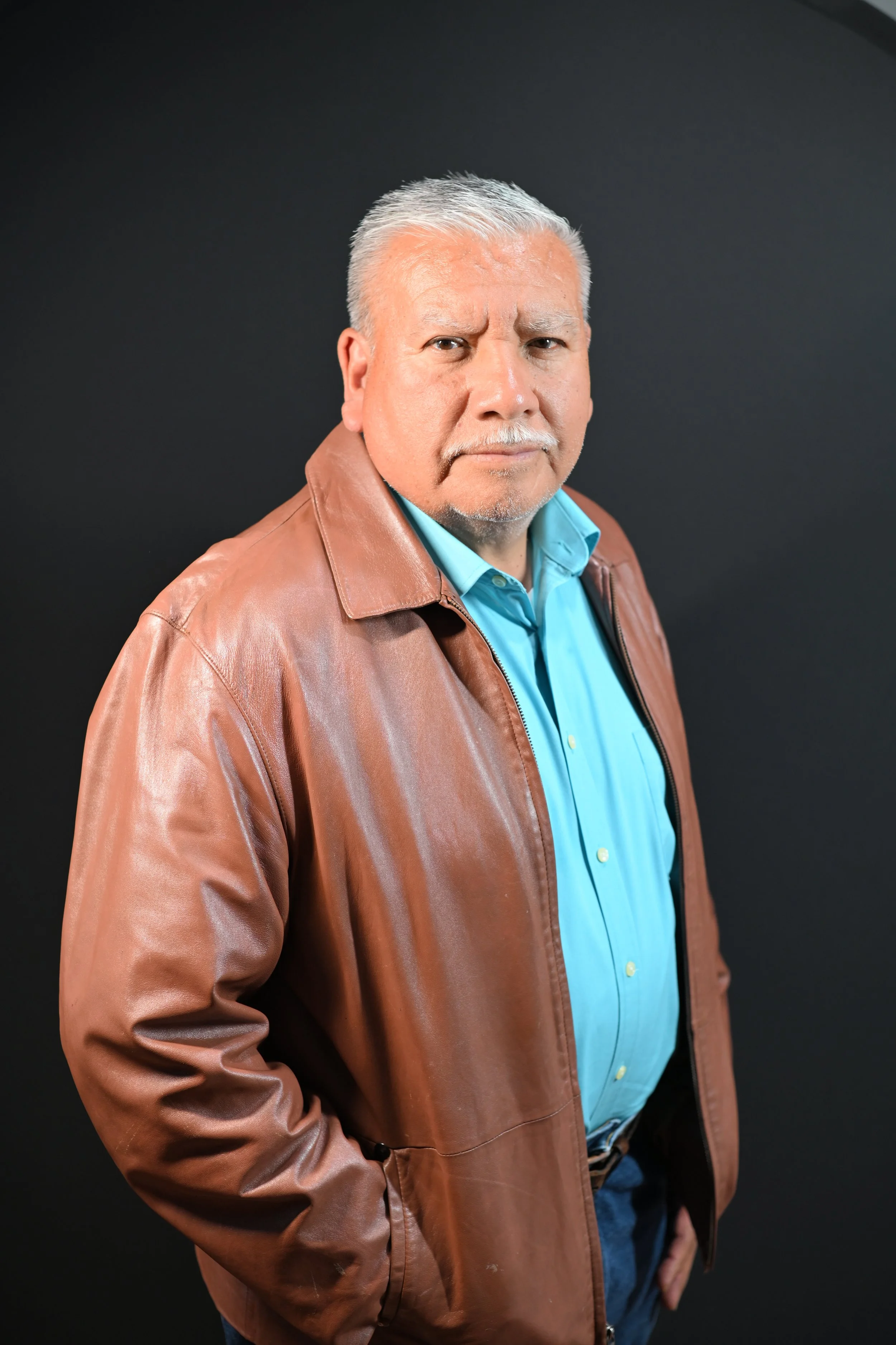A man with gray hair and a goatee wearing a blue shirt and brown leather jacket, standing against a dark background.