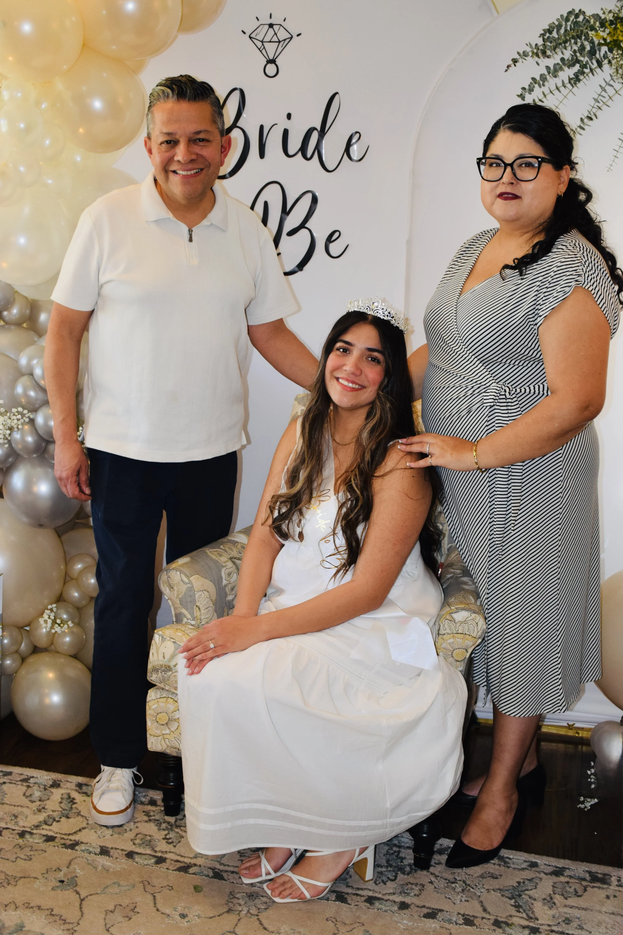 A woman in a white dress with a crown sitting on a floral patterned chair, surrounded by three adults, two women and a man, at a celebration with balloons and a sign that says 'Bride To Be'.