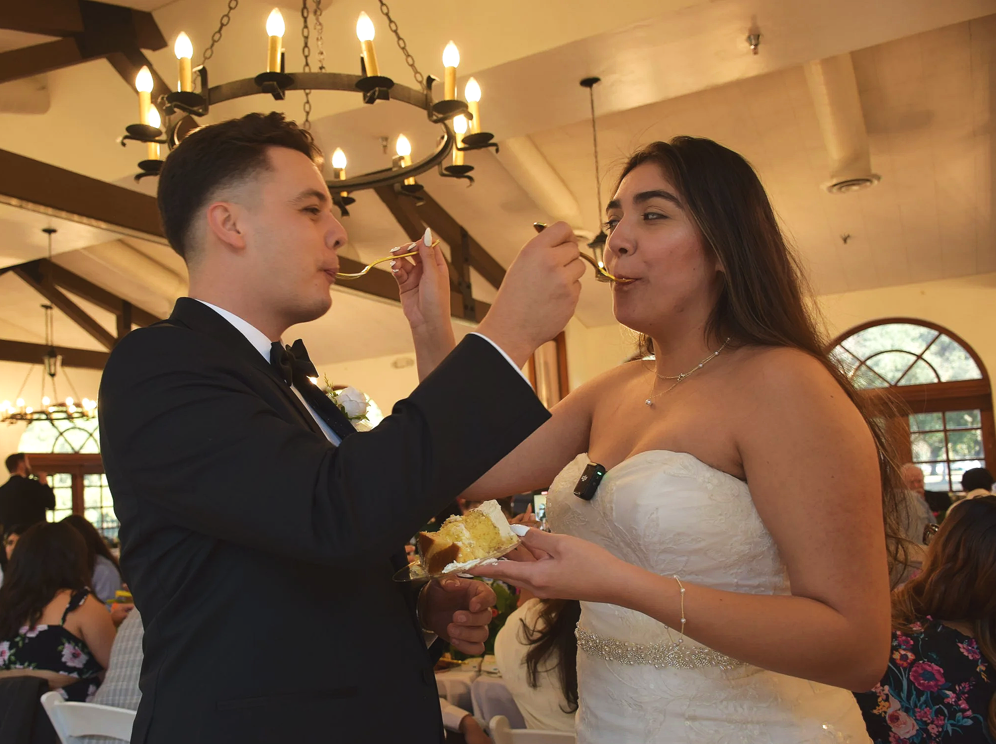 A bride and groom sharing a moment at their wedding reception while feeding each other cake. The groom is in a black tuxedo, and the bride is in a white strapless wedding gown. The setting features warm lighting, including a large, circular chandelier hanging from the ceiling.