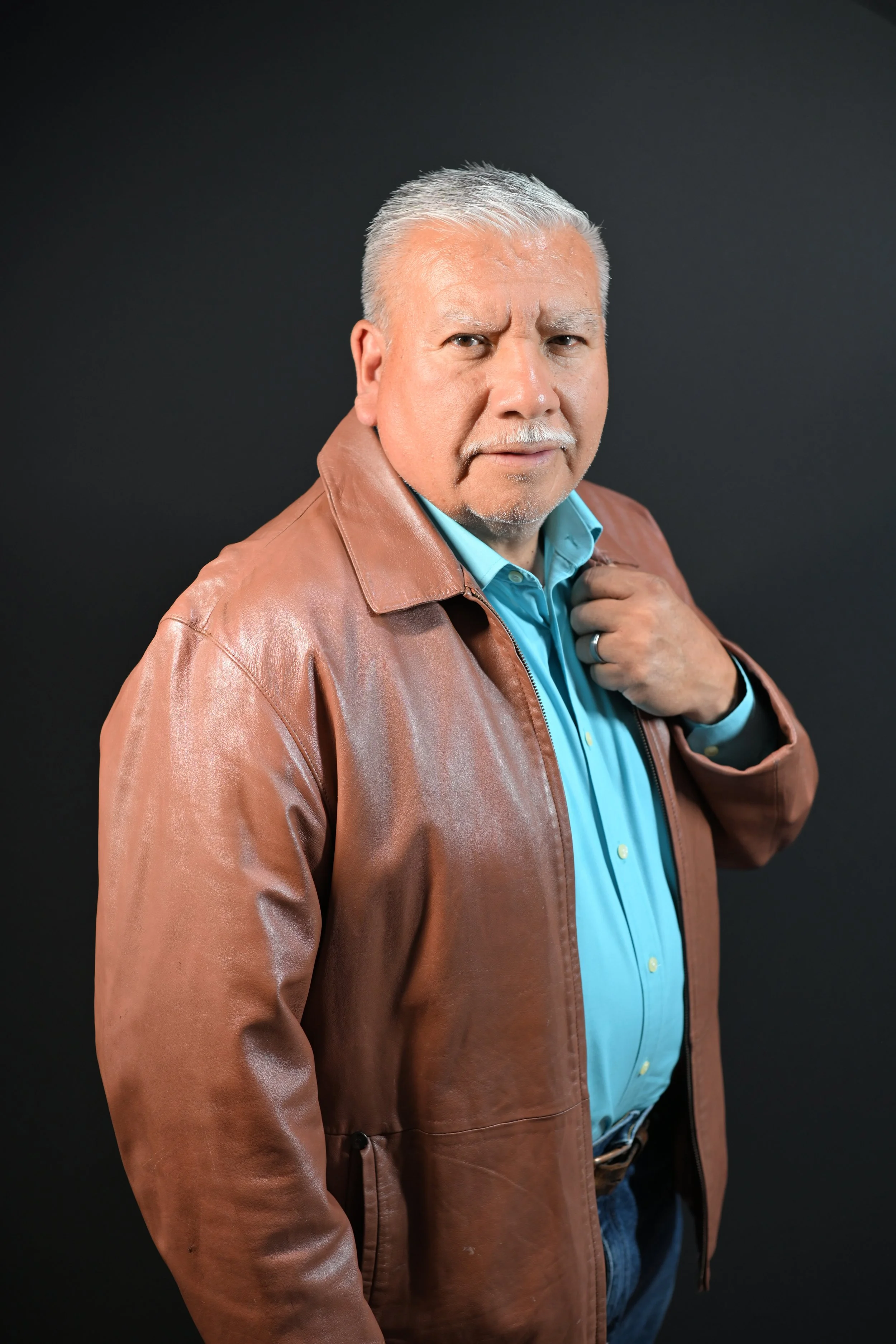 A middle-aged man with gray hair and a beard, wearing a brown leather jacket over a blue button-up shirt, standing against a black background.