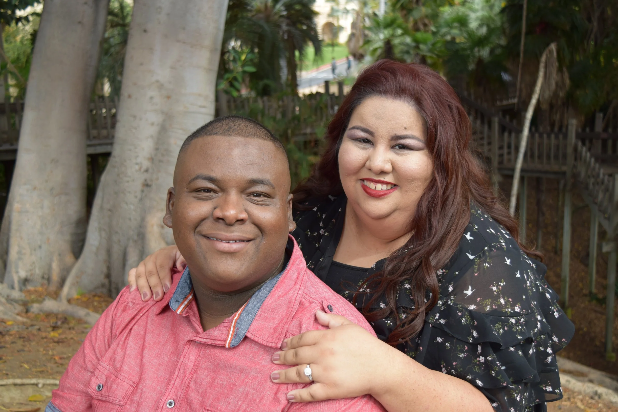 A smiling couple outdoors in a park with trees and wooden pathways. The man in the foreground is wearing a pink shirt, and the woman behind him has auburn hair and a dark floral dress.