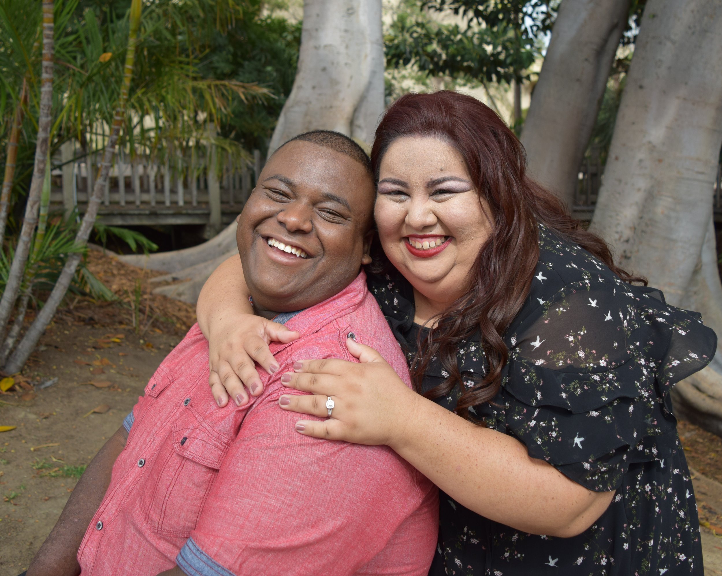 Two women embracing each other, smiling, with a background of trees and a wooden fence.