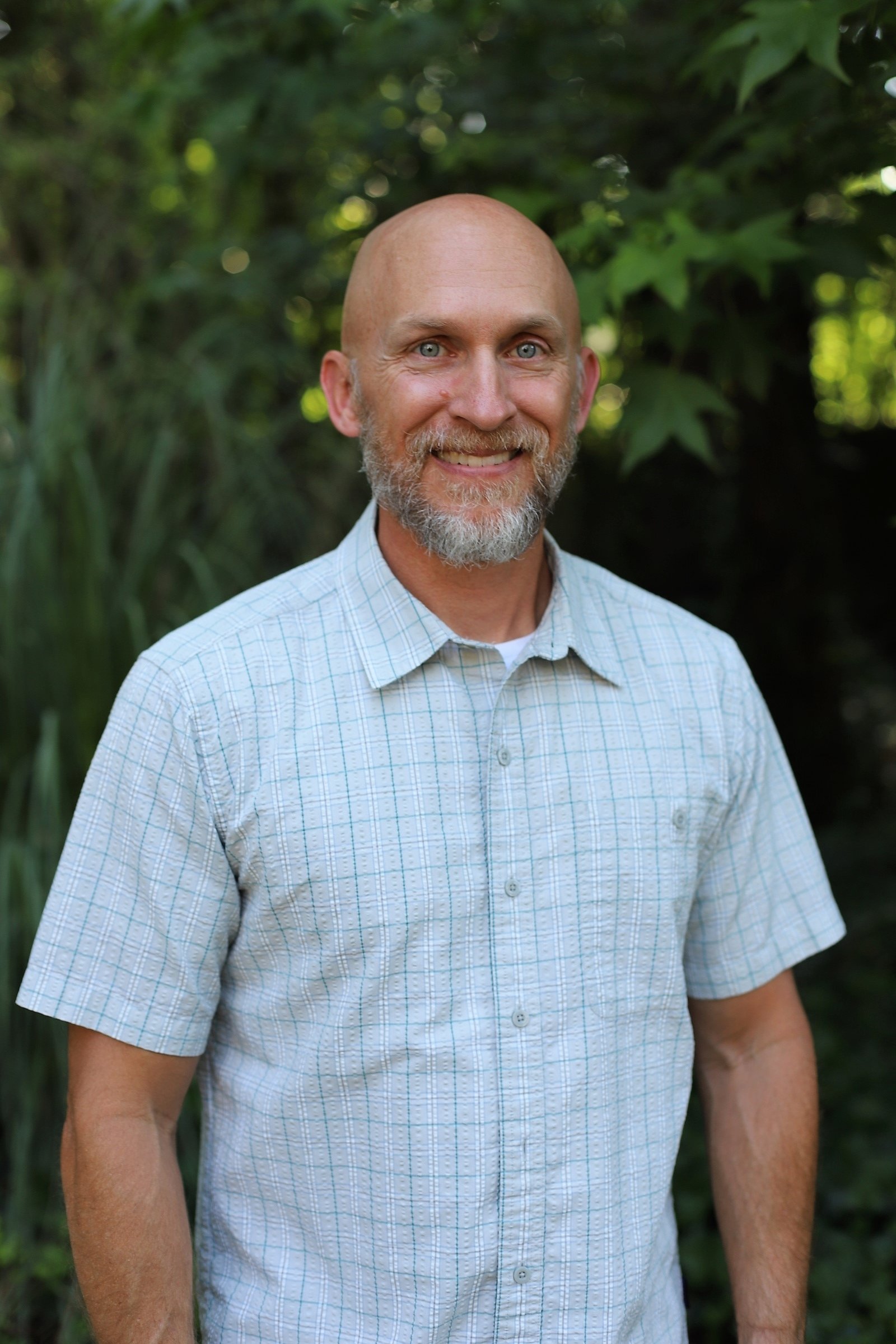 Close-up of a smiling middle-aged man with a beard and bald head, wearing a light-colored, checkered short-sleeve shirt, standing outdoors with green foliage in the background.