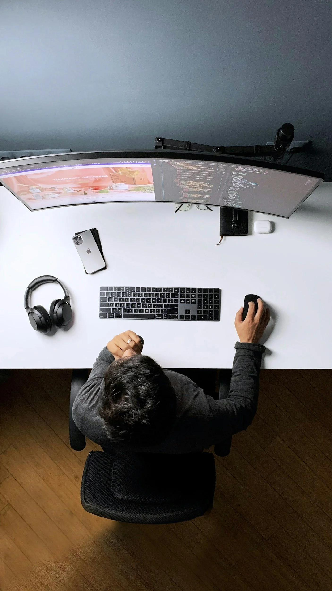 Overhead view of a person working at a white desk with dual curved monitors, a keyboard, a mouse, headphones, and a smartphone.