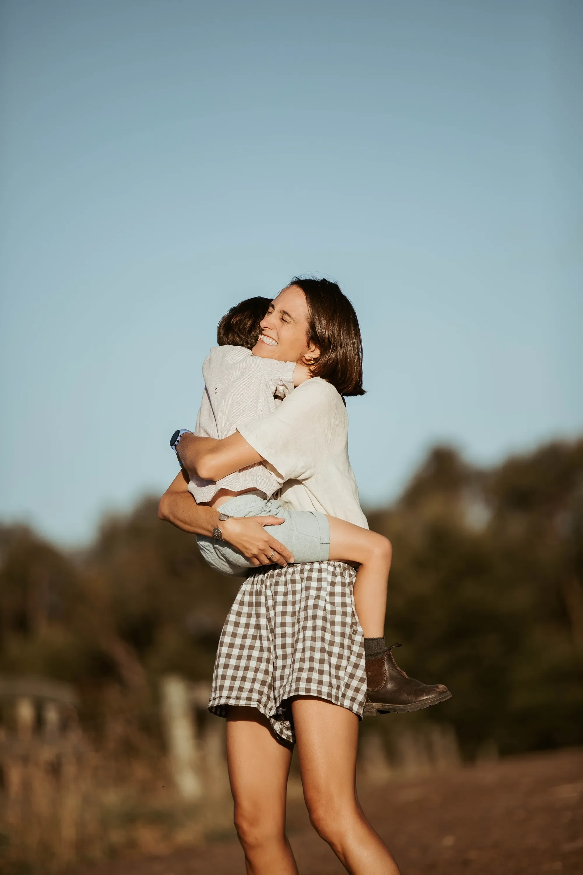 A woman with short brown hair wearing checkered shorts and brown boots is holding a young child in her arms. The child is hugging her, and they are both smiling. The background features a clear blue sky and blurred nature scenery.