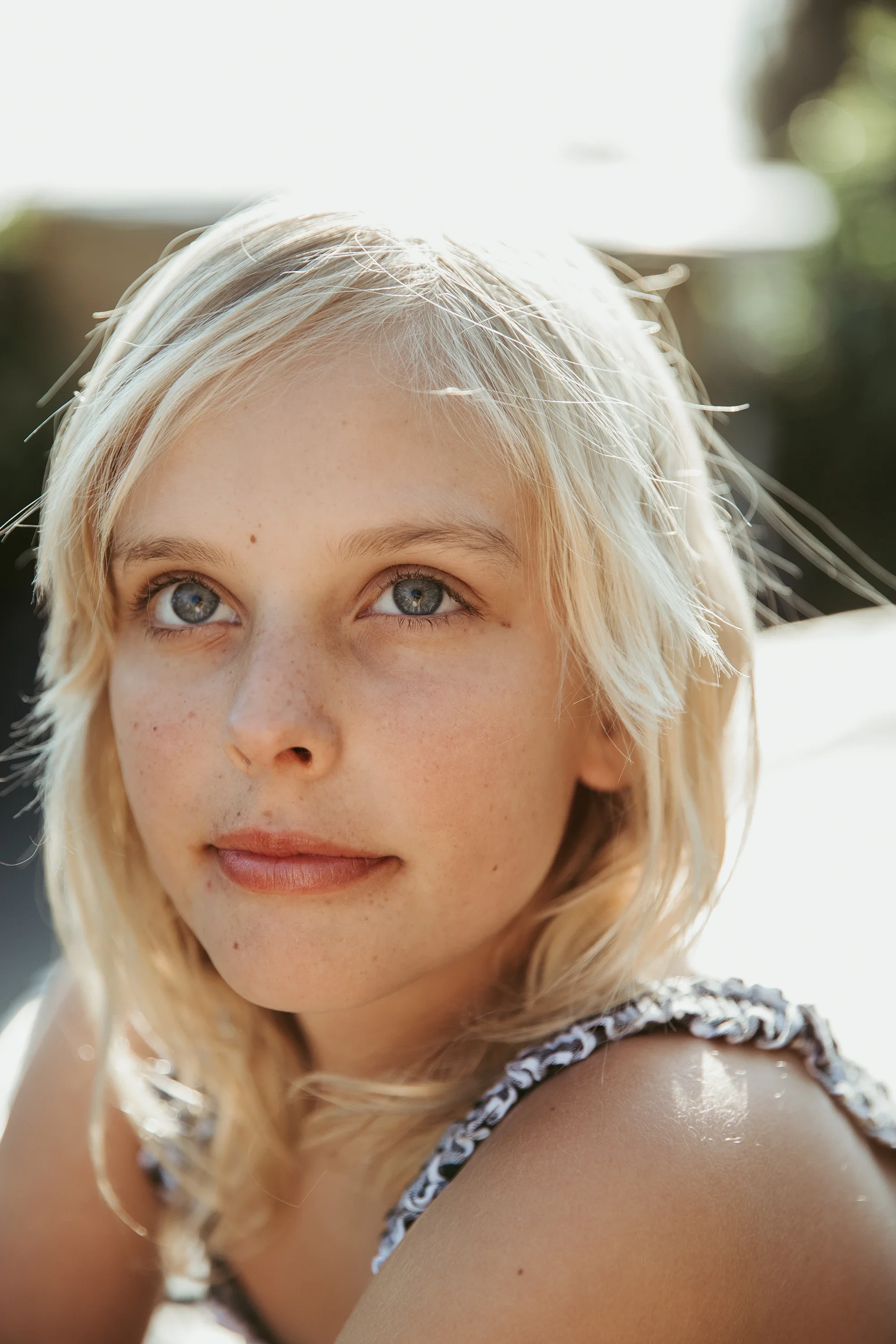 Close-up of a young woman with blonde hair, blue eyes, and light skin, outdoors with sunlight highlighting her hair and face, wearing a patterned sleeveless top.