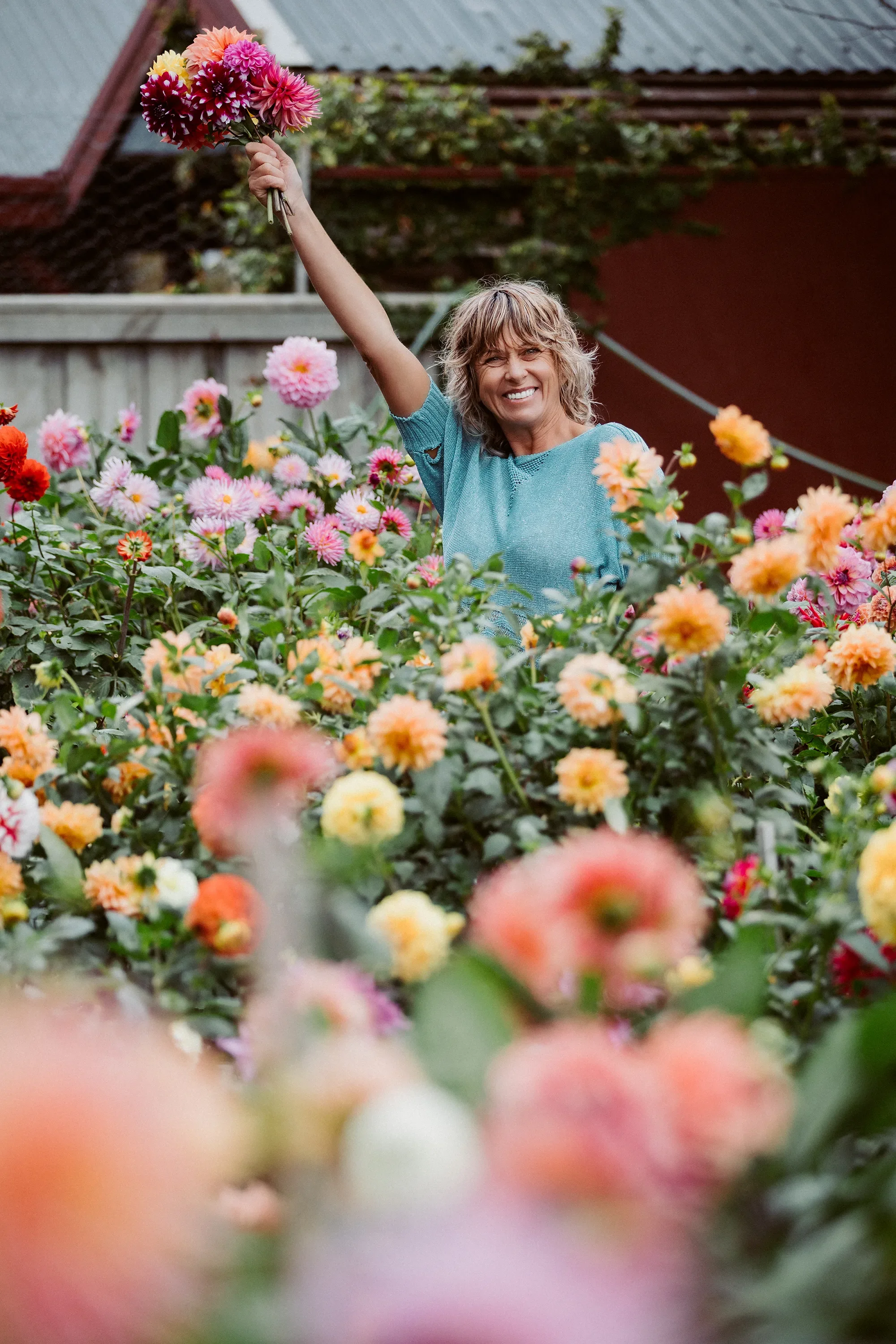 A woman smiling and holding a bouquet of pink, orange, and yellow flowers in a garden setting with various flowers and a shed in the background.
