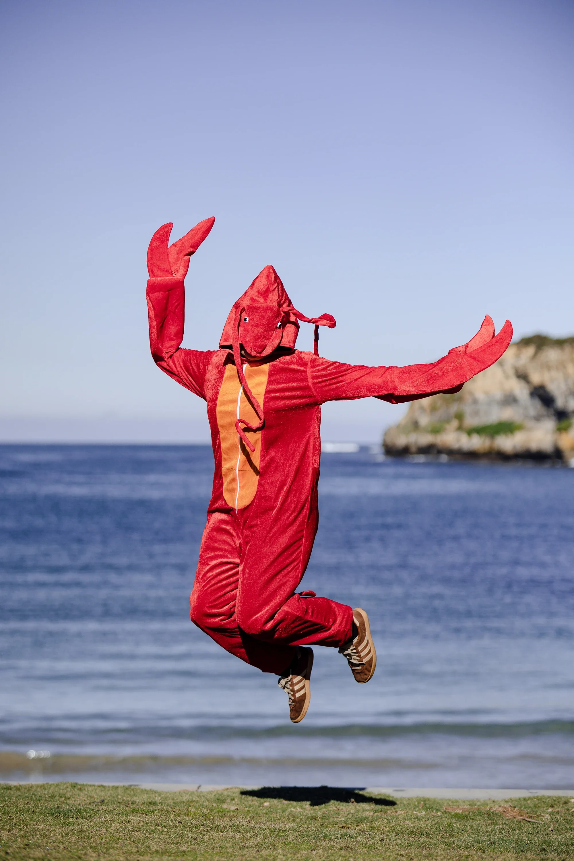 Person dressed in a lobster costume jumping in front of the ocean with cliffs in the background.