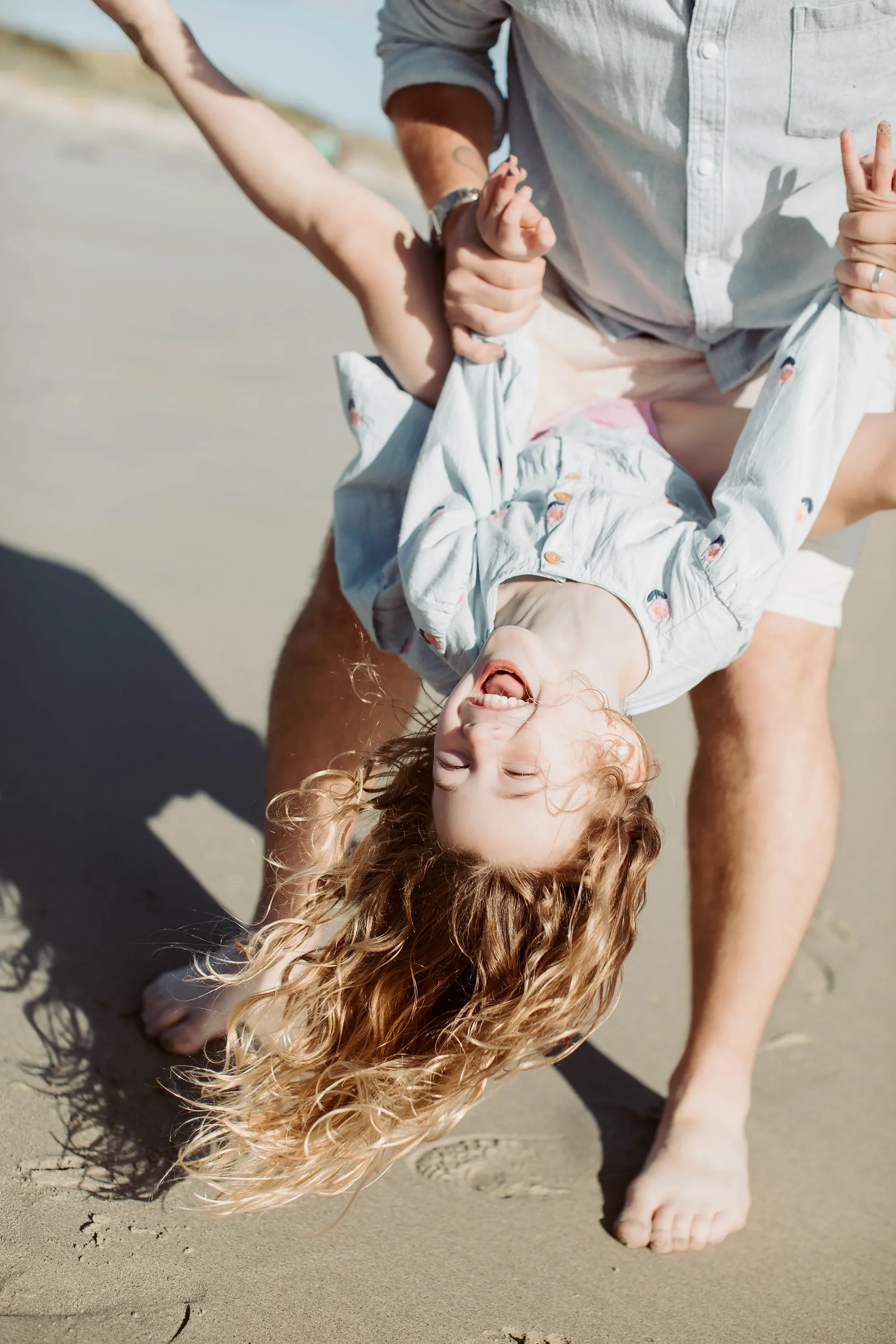 A young girl with long, curly red hair lying on a sandy beach, smiling, with a person holding her hand and foot. The girl is wearing a light-colored shirt with a colorful pattern.
