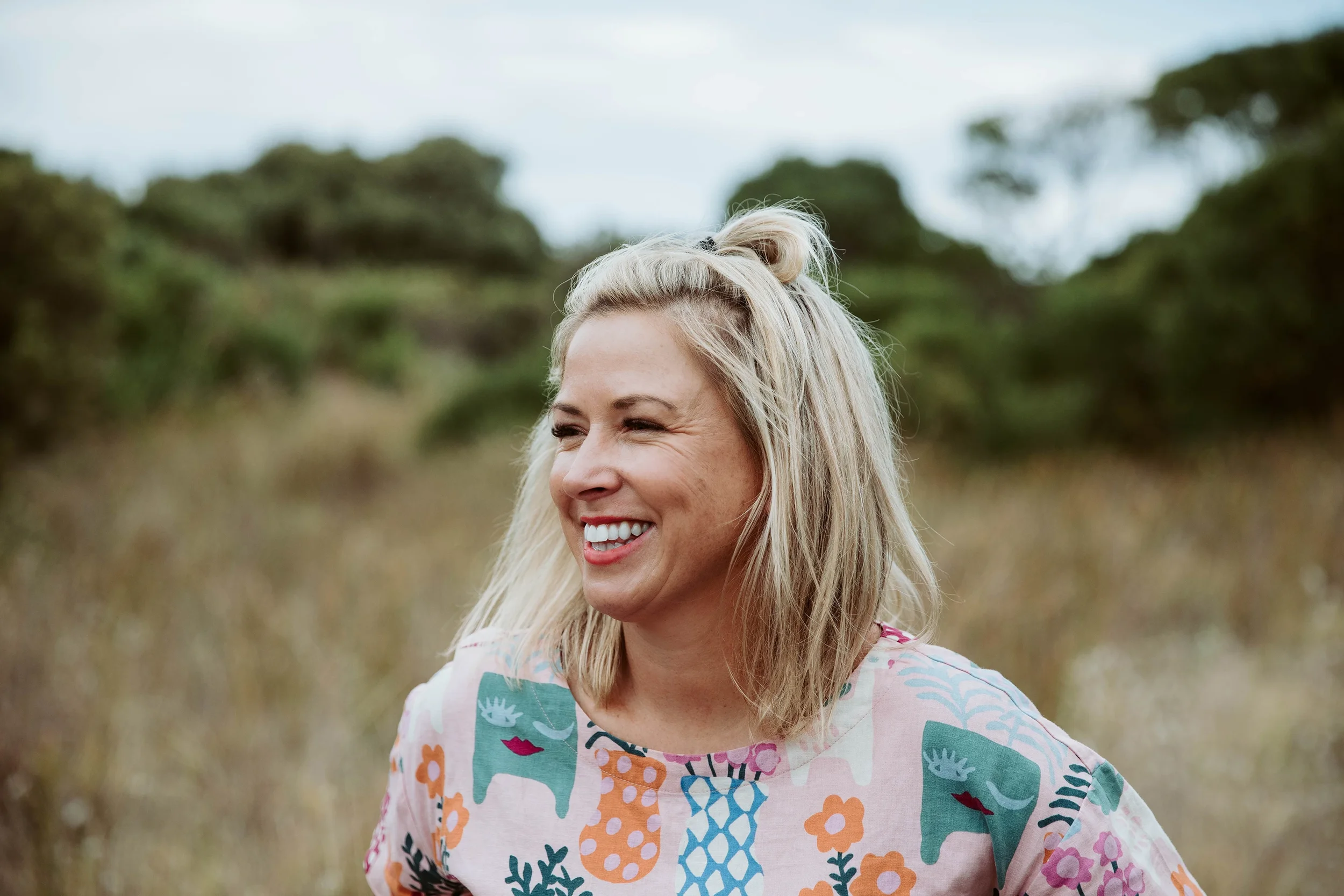 A woman with long blonde hair smiling outdoors on a cloudy day, wearing a colorful patterned top with abstract shapes and nature motifs, in a natural setting with trees and grass in the background.