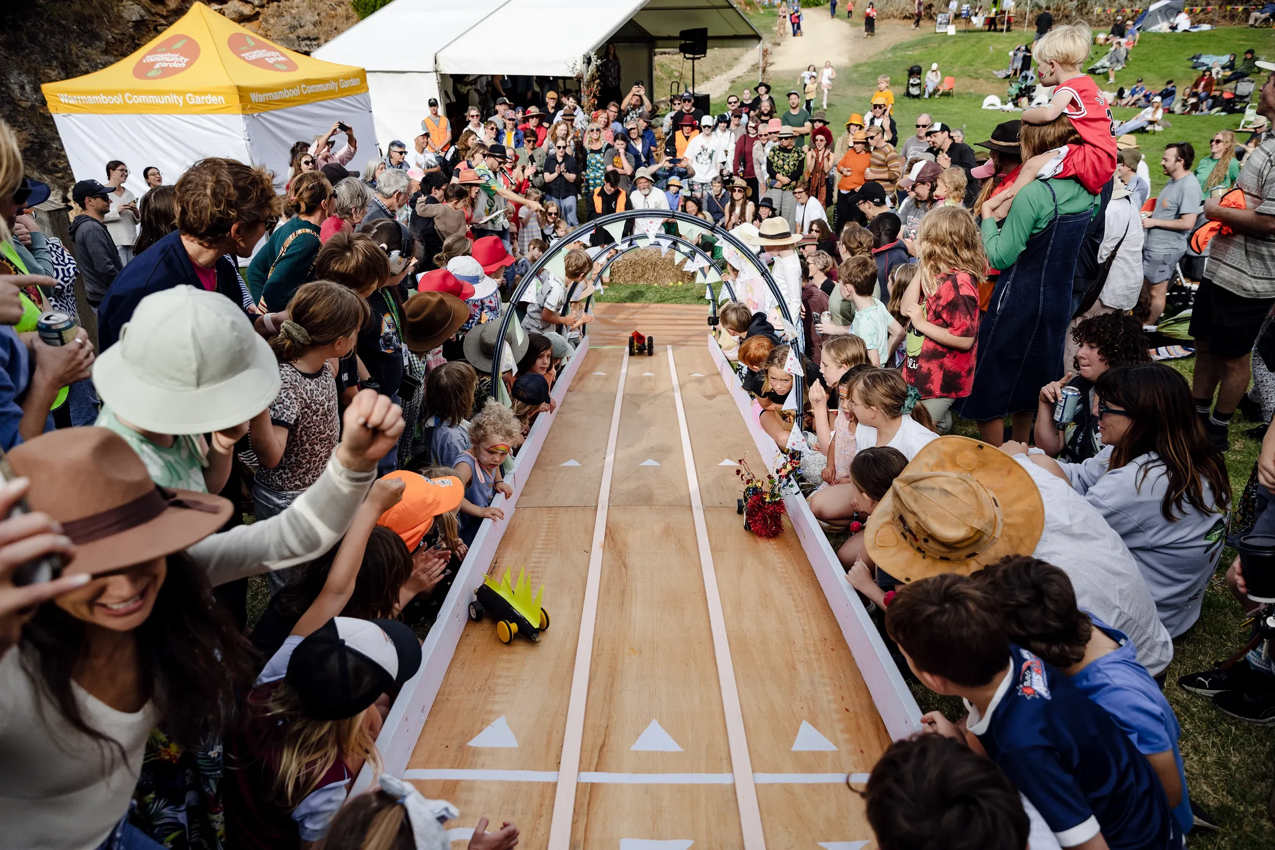 Crowd of people gathered around a wooden race track with small cars, at an outdoor festival. Children and adults watch the race, some wearing hats and sunglasses, with a large white event tent and a yellow banner reading 'Warmambool Community Garden' in the background.