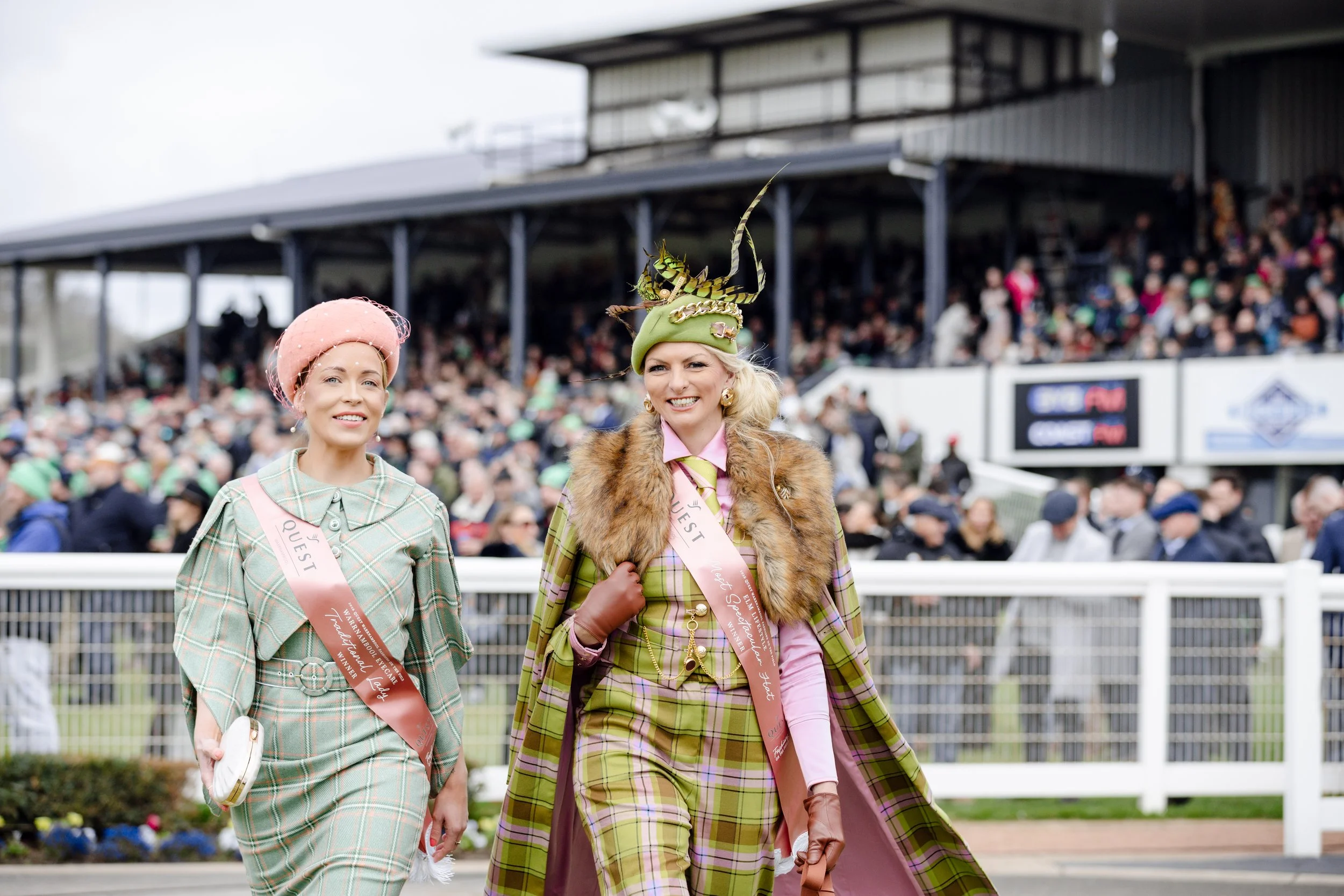 Two women dressed in colorful, vintage-style outfits walking at a racetrack, with a large crowd of spectators in the background.