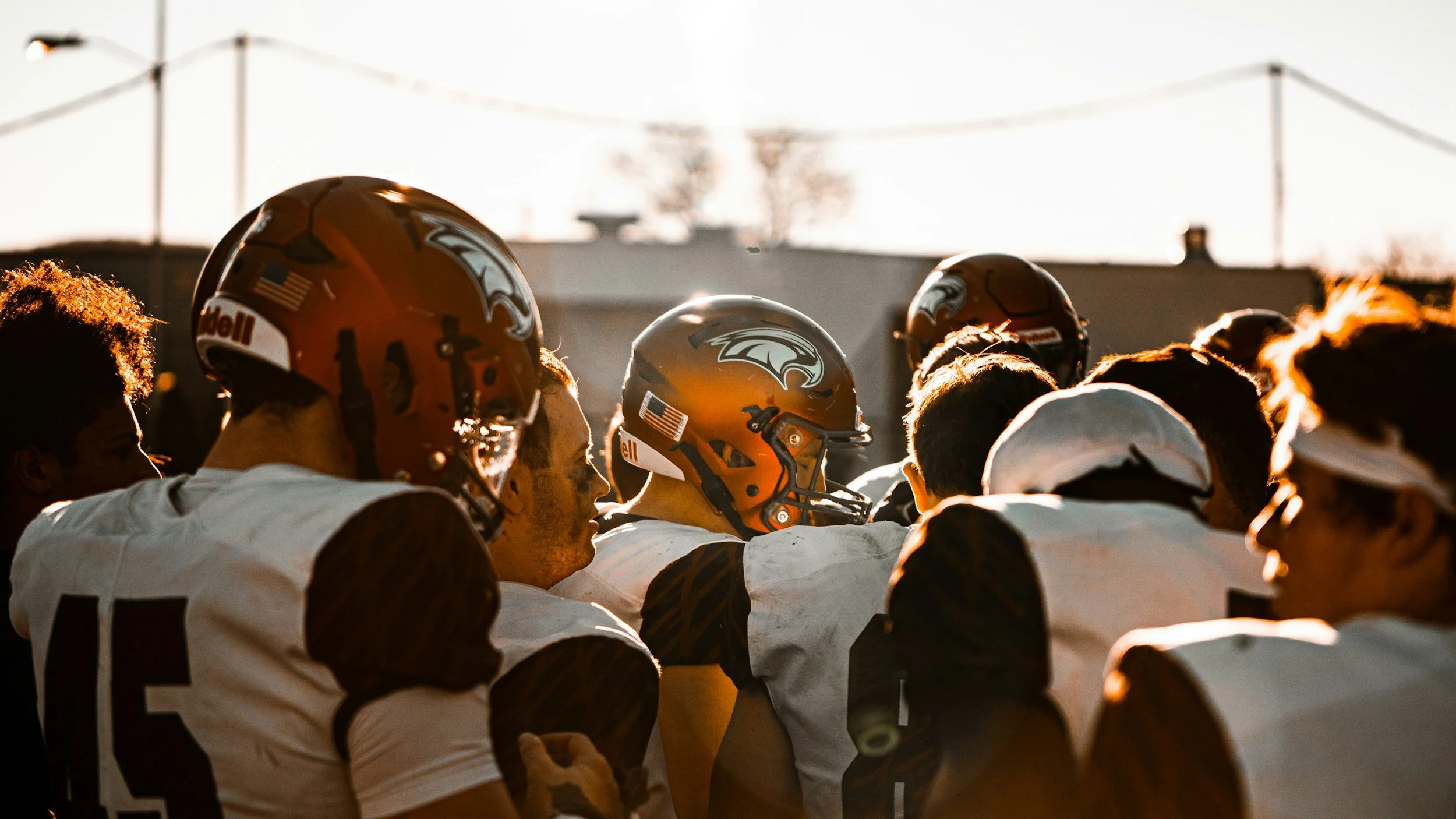 A group of football players wearing helmets and white uniforms, standing close together during sunset or sunrise.