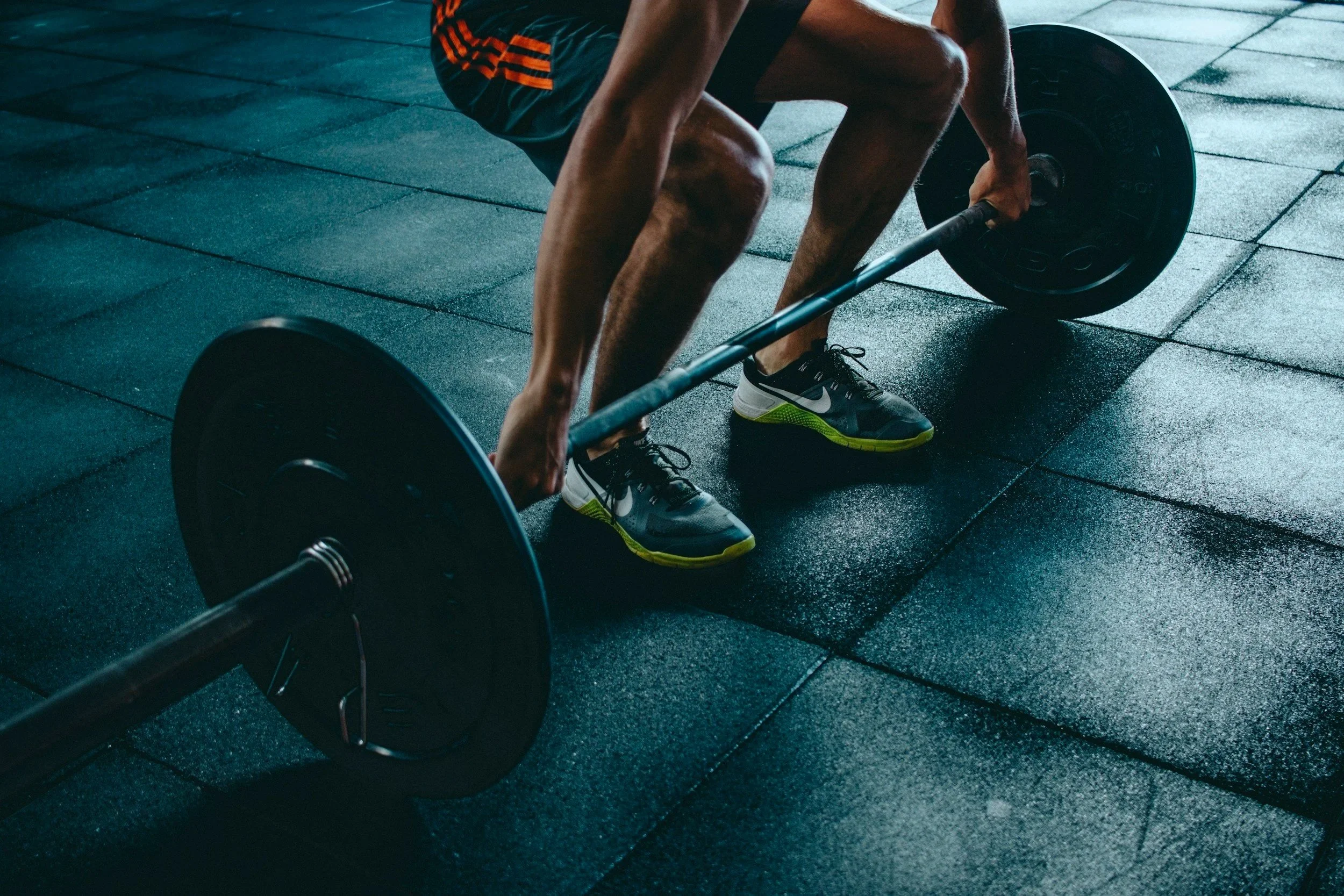 A person in athletic shorts and Nike sneakers is bent over, lifting a barbell with weights on a gym floor.