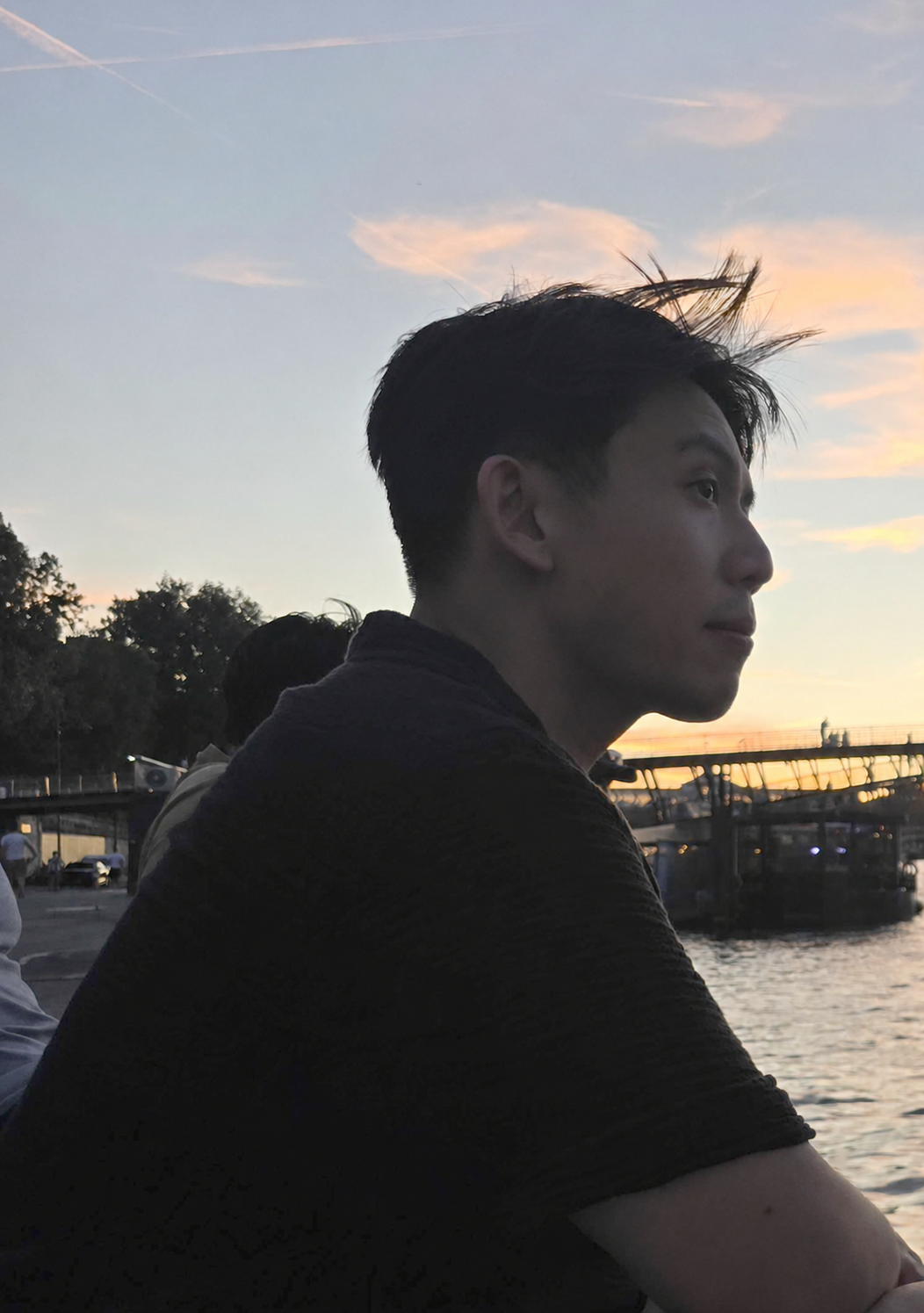 A young man with styled hair sitting near a body of water during sunset.