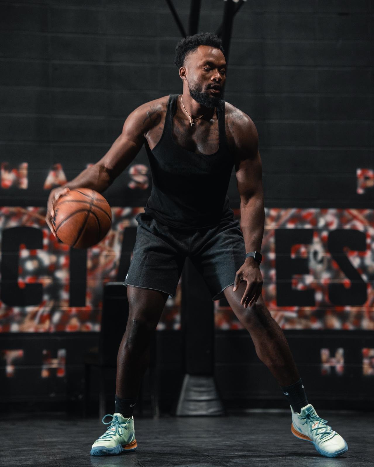A man wearing a black tank top and black shorts dribbling a basketball on an indoor court.