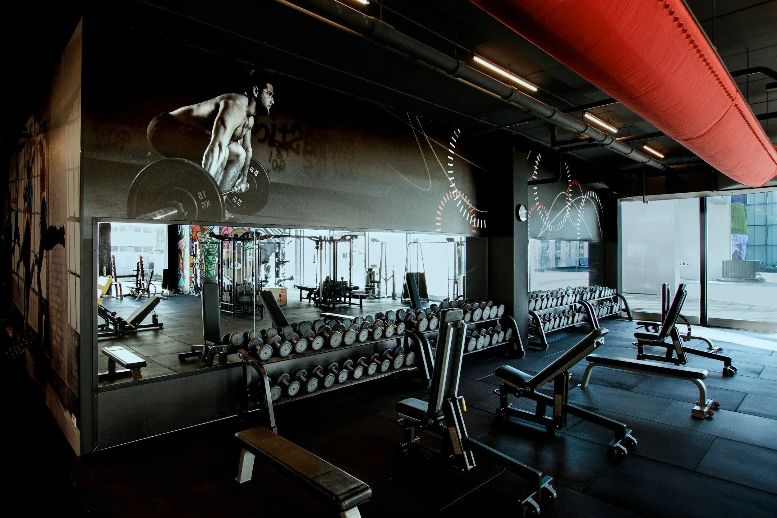 Inside a modern gym with workout equipment, dumbbells, benches, and exercise machines. A large black-and-white mural of a man lifting weights is on the wall, and there are large windows allowing natural light to enter the space.