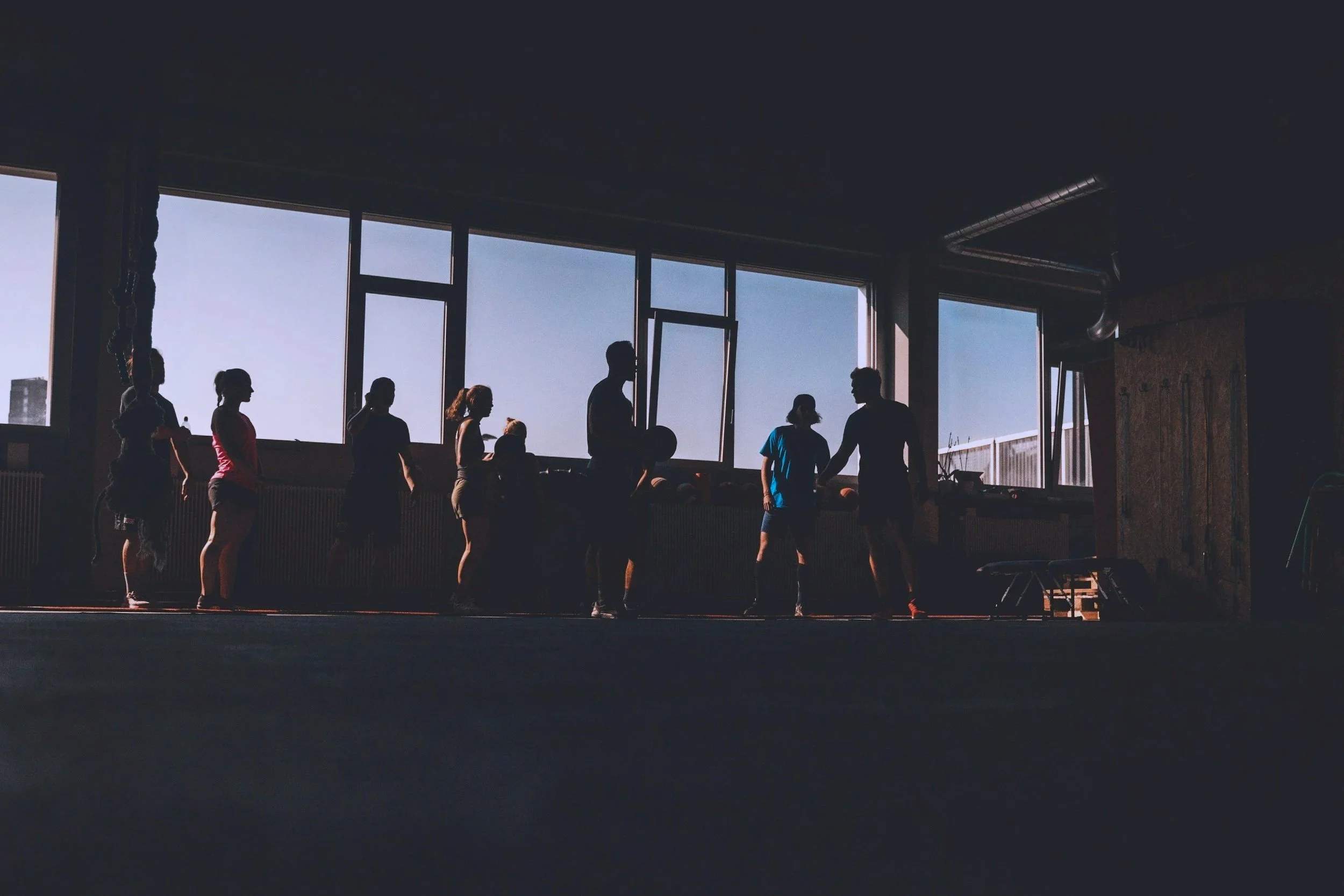 Silhouettes of people engaging in a fitness or dance class inside a large studio with floor-to-ceiling windows showing a clear sky outside.