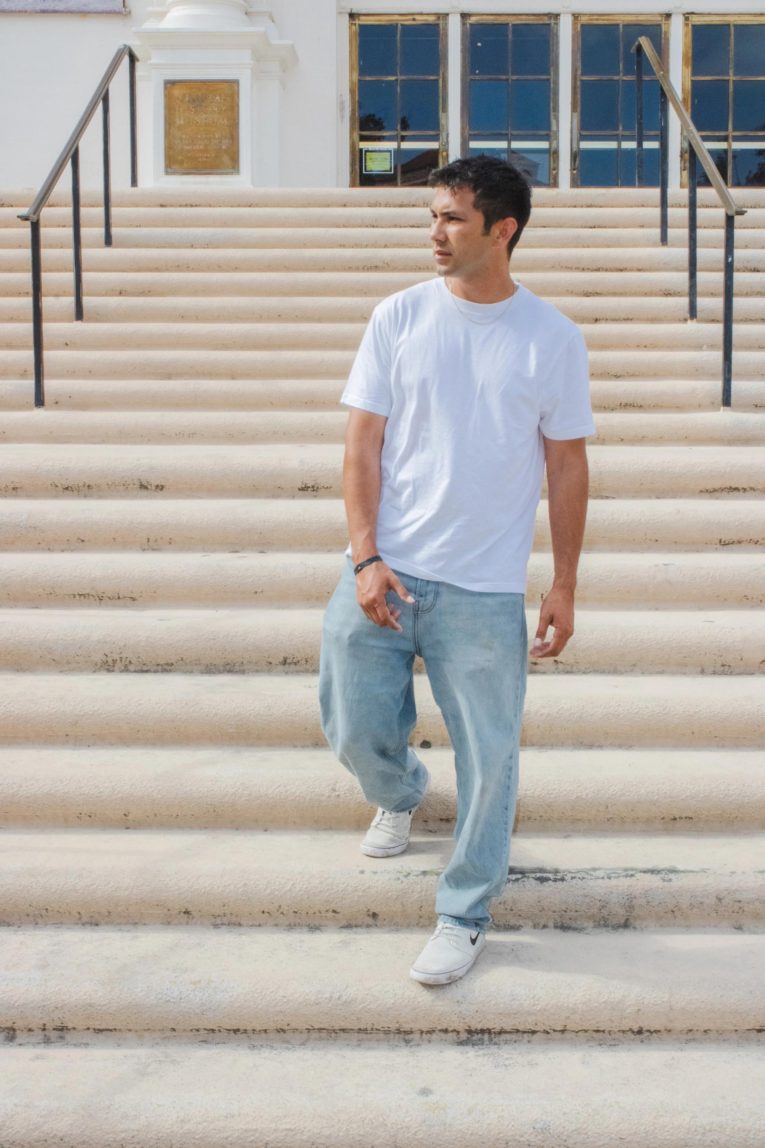 A man wearing a white t-shirt, light blue jeans, and white sneakers walking down white steps outside a building with large windows and a plaque.