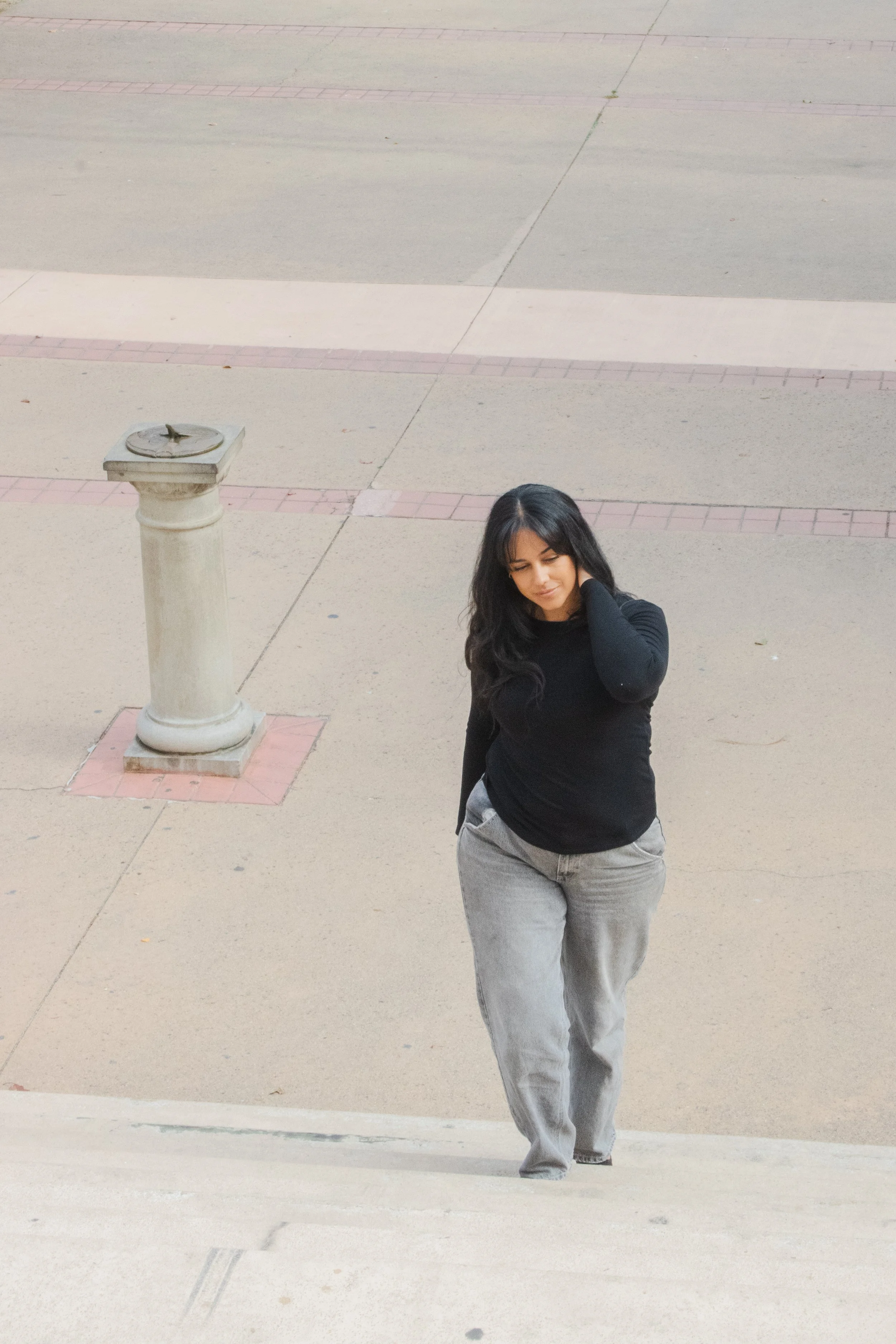 A young woman with long black hair wearing a black long sleeve shirt and gray jeans walking on a sidewalk. She is touching her neck with her right hand and looking down with a slight smile. The background shows an empty paved area with a white cylindrical post.