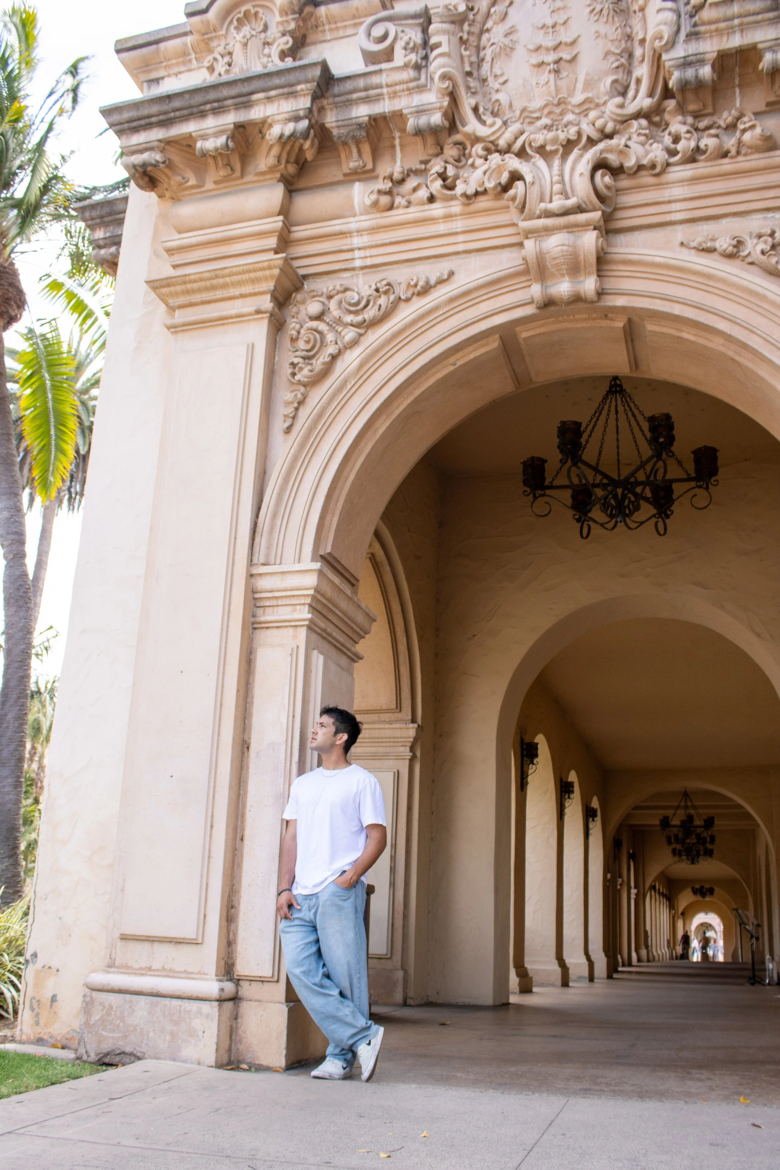 A young man in a white t-shirt and light blue jeans stands with his back leaning against a large stone pillar in a corridor with classical architectural details, including ornate arches, a chandelier, and lantern-style wall sconces, with palm trees visible in the background.