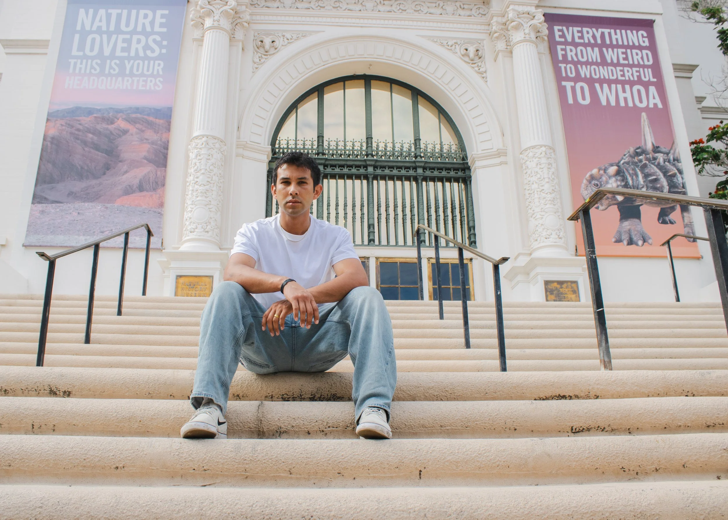 A young man sitting on the steps of an ornate building with white columns and decorative arches. Two large banners hang on either side of the entrance, one featuring a mountain landscape and the other a dinosaur, with texts about nature and curiosity. The young man wears a white t-shirt, light jeans, and white sneakers, with a serious expression.