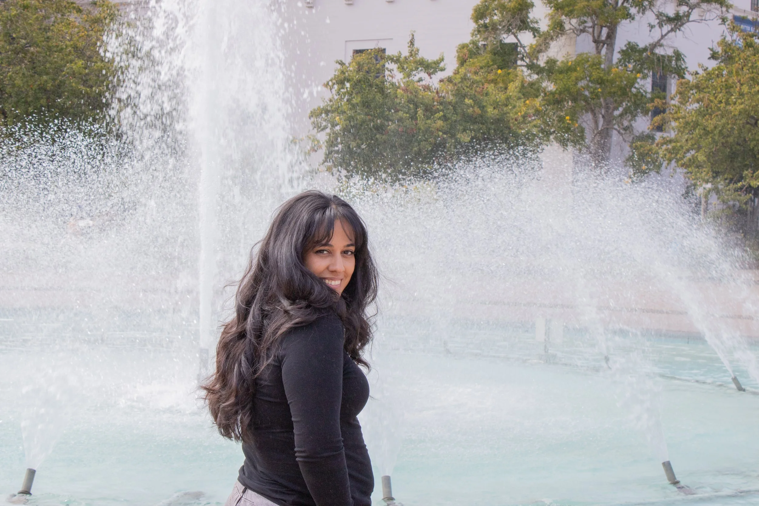 A young woman with long dark hair smiling and standing in front of a water fountain, with trees and a building in the background.