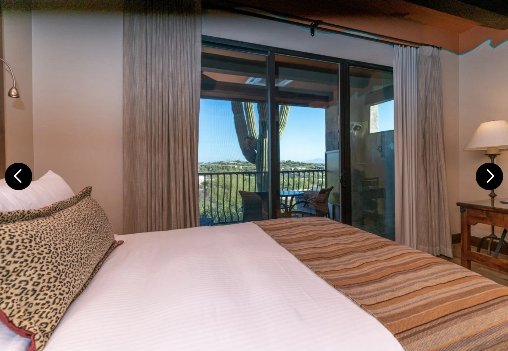 A bedroom with a bed, leopard print pillow, and striped bed runner, facing a balcony with patio table and chairs, view of green landscape and cactus outside.