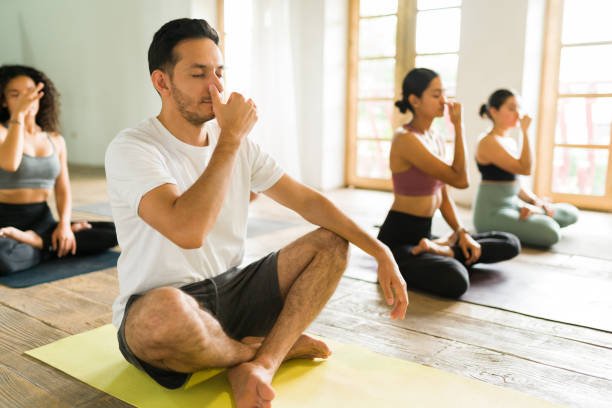 A group of people practicing yoga in a bright studio with large windows, sitting cross-legged with hands in prayer position, eyes closed, focusing inward.