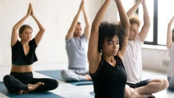 Group of people practicing yoga and meditation in a bright studio with large windows.