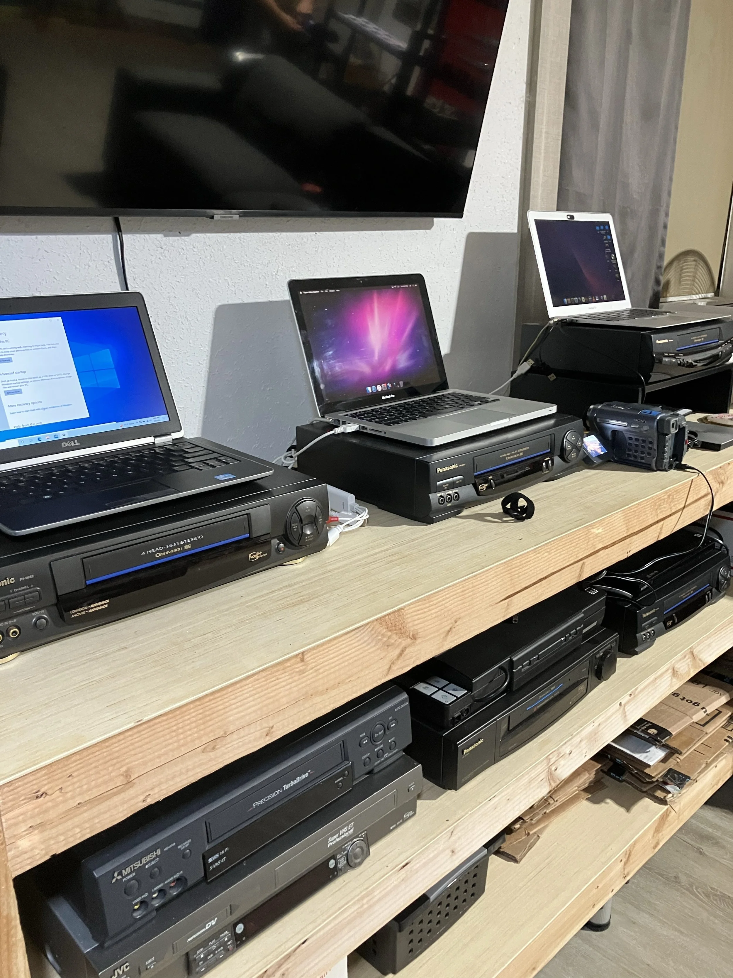 A variety of electronic devices including laptops, a DVD/VHS player, and other media equipment are arranged on a wooden shelf in this image.