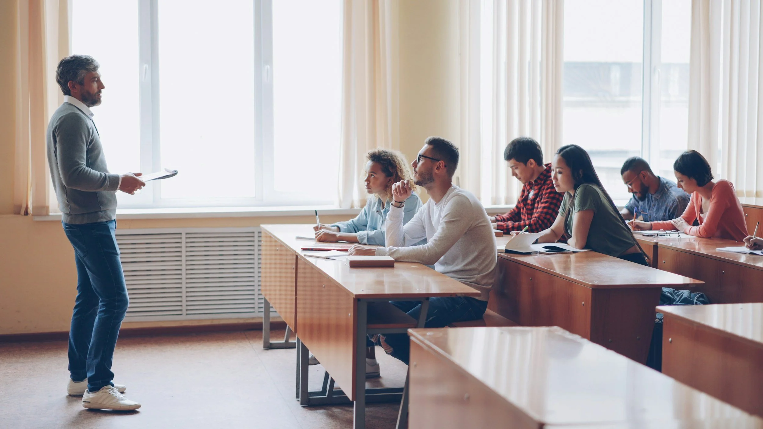 A classroom scene with a teacher standing at the front and students sitting at desks, listening attentively.