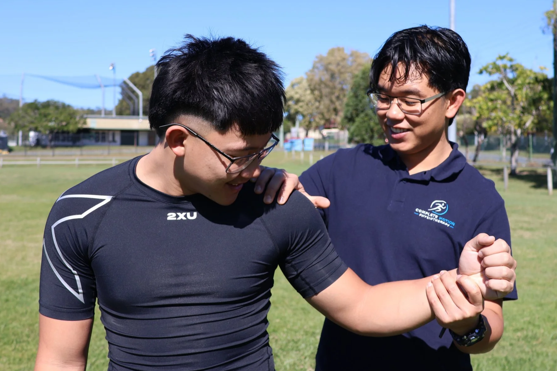 Two young men, both wearing glasses, are outdoors on a grass field on a sunny day. One man is wearing a black athletic shirt with white accents, and the other is in a navy blue shirt with a logo. The man in black is showing his arm muscles to the man in navy blue, who is smiling and holding his shoulder.