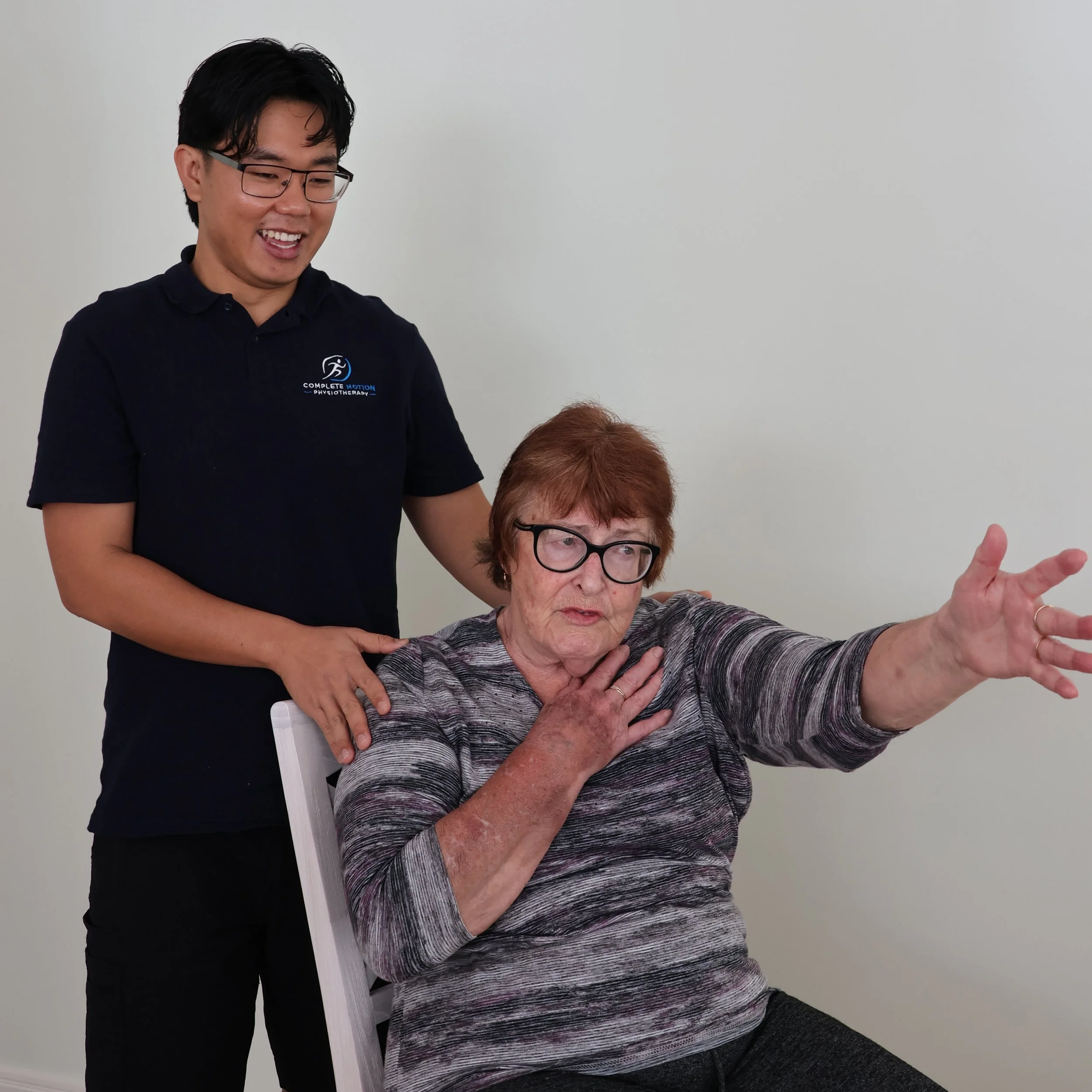 A healthcare professional standing next to an elderly woman sitting in a chair, performing a stretching exercise with her arm. The woman is wearing glasses and a striped shirt, while the professional is wearing a black polo shirt with a logo. They are in a plain room with a white wall.