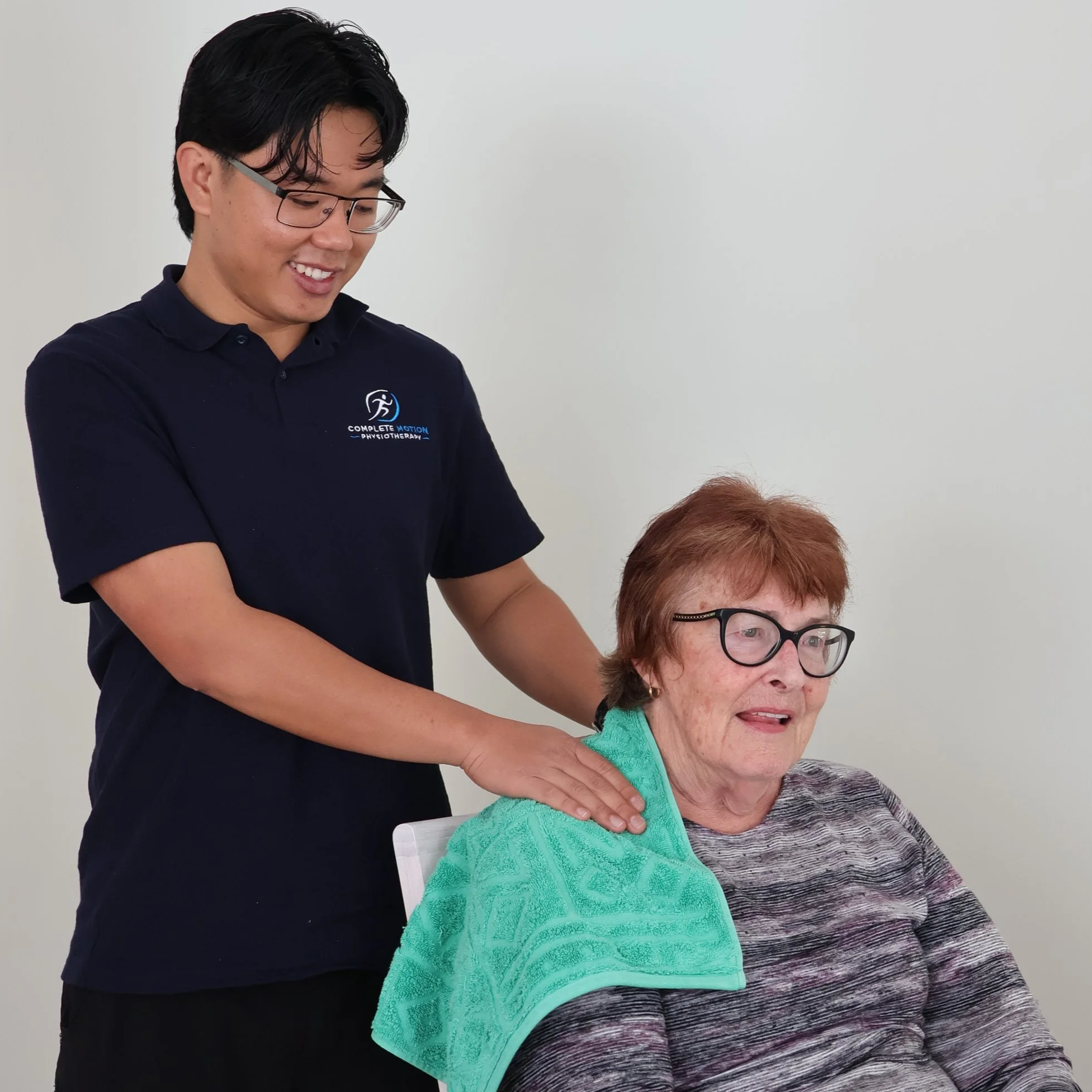 A physical therapist wearing glasses and a navy polo shirt with a logo on it is smiling as she massages the shoulder of an elderly woman wearing glasses and a striped sweater. The woman is seated, and a green towel is draped over her shoulders.