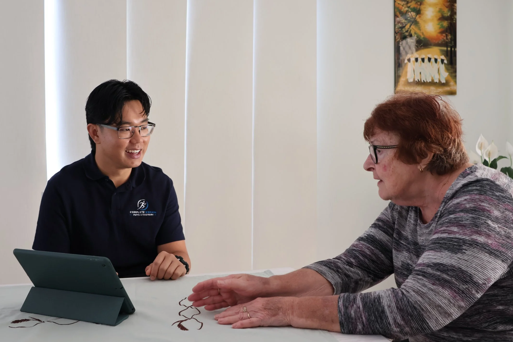 A young man and an elderly woman sit across from each other at a table, engaging in a conversation. The young man is smiling, wearing glasses and a collared shirt, with a tablet device in front of him. The elderly woman has reddish-brown hair, glasses, and is wearing a patterned sweater with her right hand extended, possibly demonstrating a hand exercise or activity. A tablet is also on the table, propped up with a cover. The background features vertical blinds and a colorful painting on the wall.