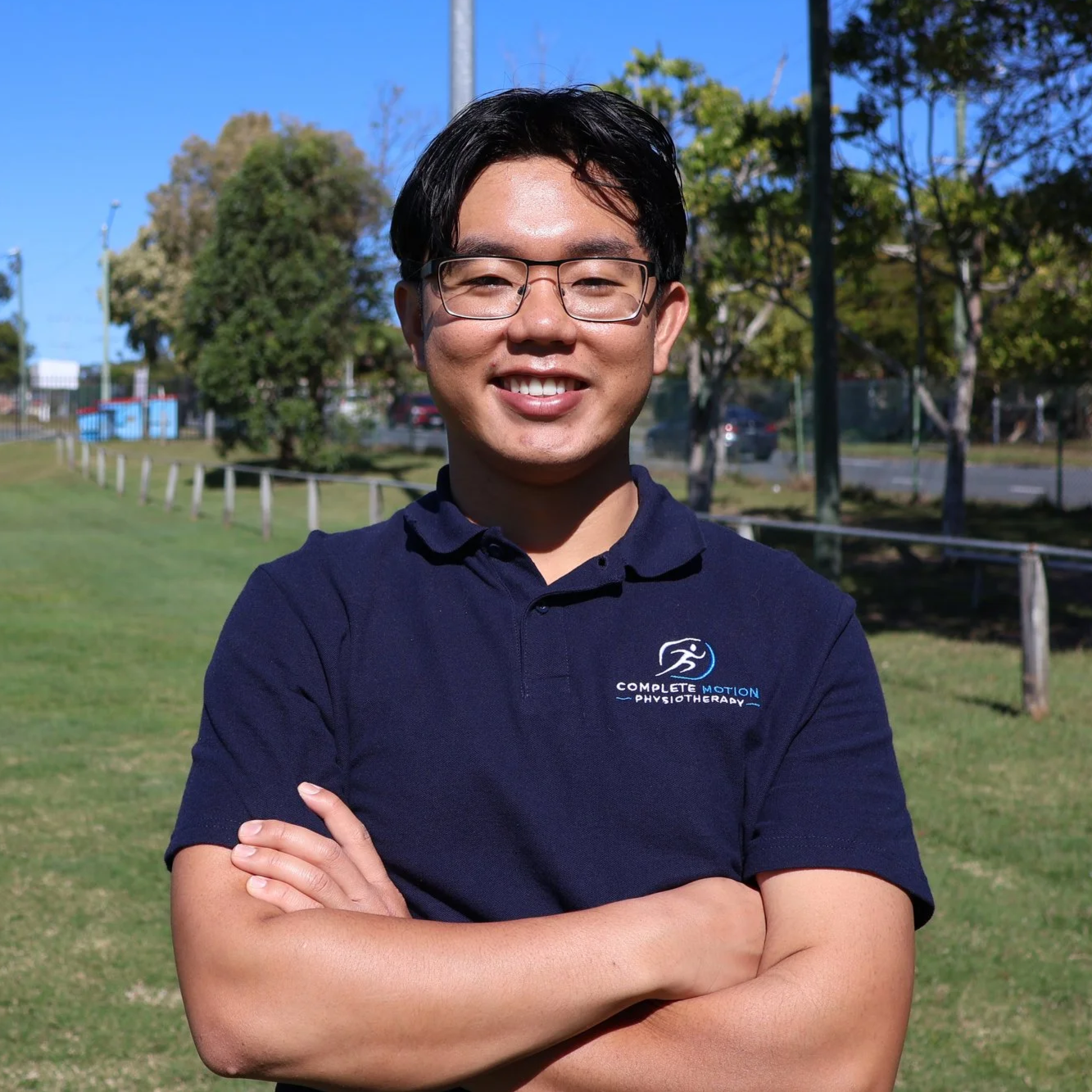 A young man with glasses smiling, crossing his arms while standing outdoors on a sunny day. Trees and a park are visible in the background. He is wearing a navy blue polo shirt with a logo that reads 'Complete Motion Physiotherapy.'