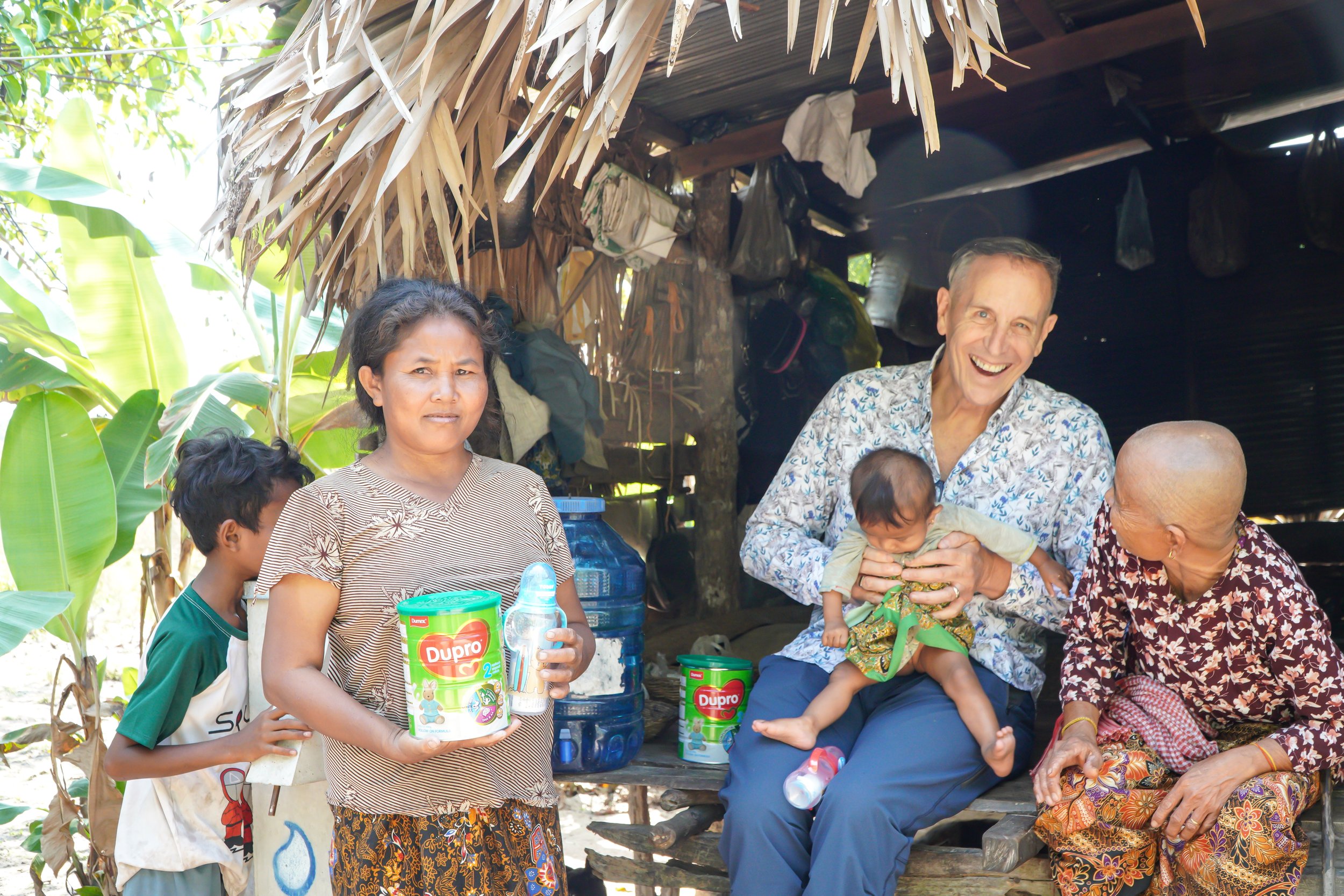 A group of people in a rural outdoor setting, with greenery and banana leaves visible. A woman in the center holding a container of Dupro baby formula, a young boy standing beside her, a smiling man holding a toddler, and an elderly woman sitting nearby. The scene appears to depict a community gathering or family celebration.