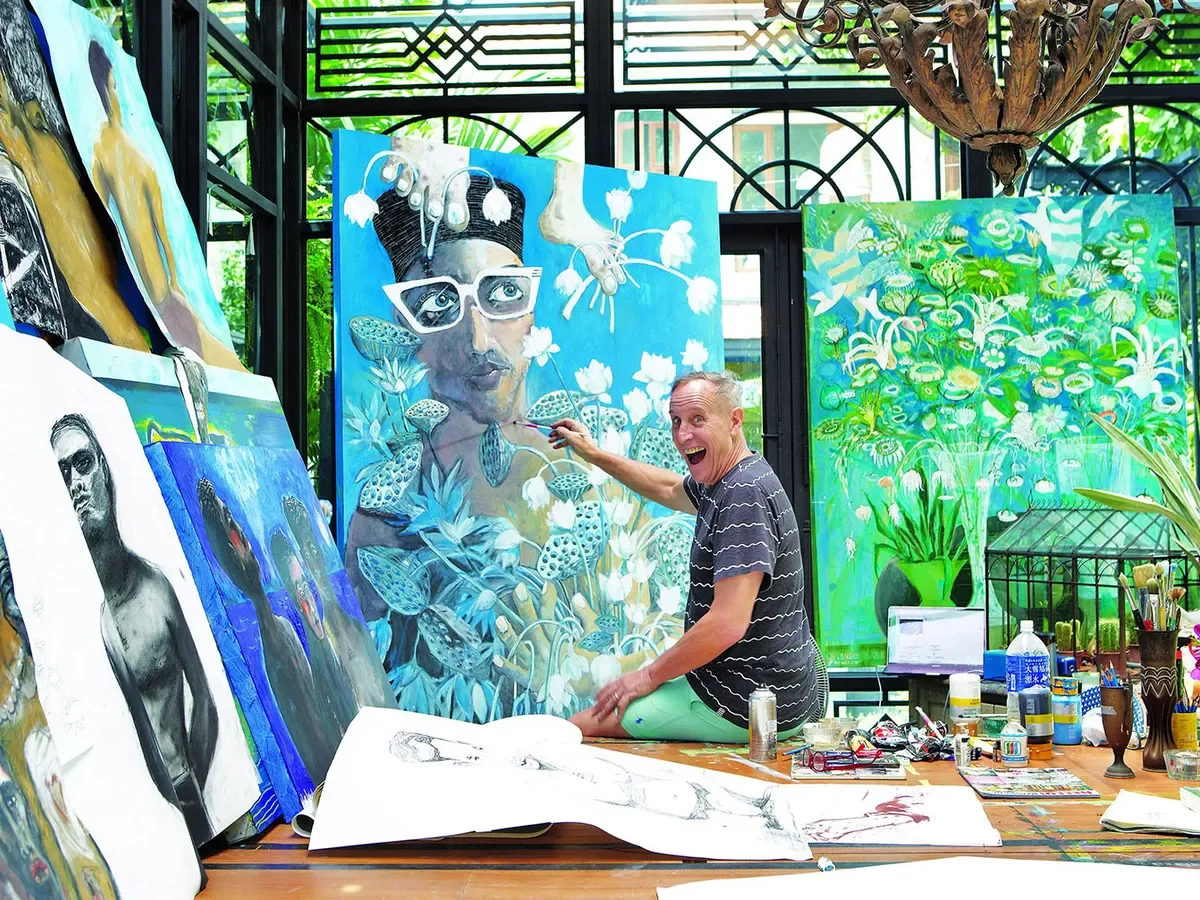 An artist painting a large portrait of a woman with glasses, surrounded by flowers, with additional artwork and art supplies on the table, in a bright, green-filled studio.