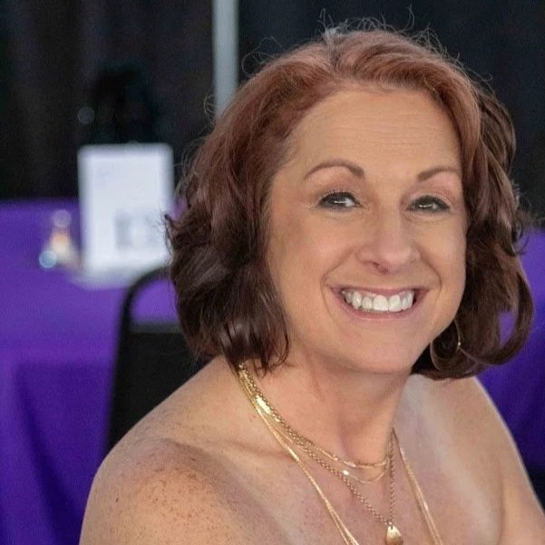 A smiling woman with shoulder-length reddish-brown hair, wearing makeup and jewelry, at an indoor event with a purple tablecloth in the background.