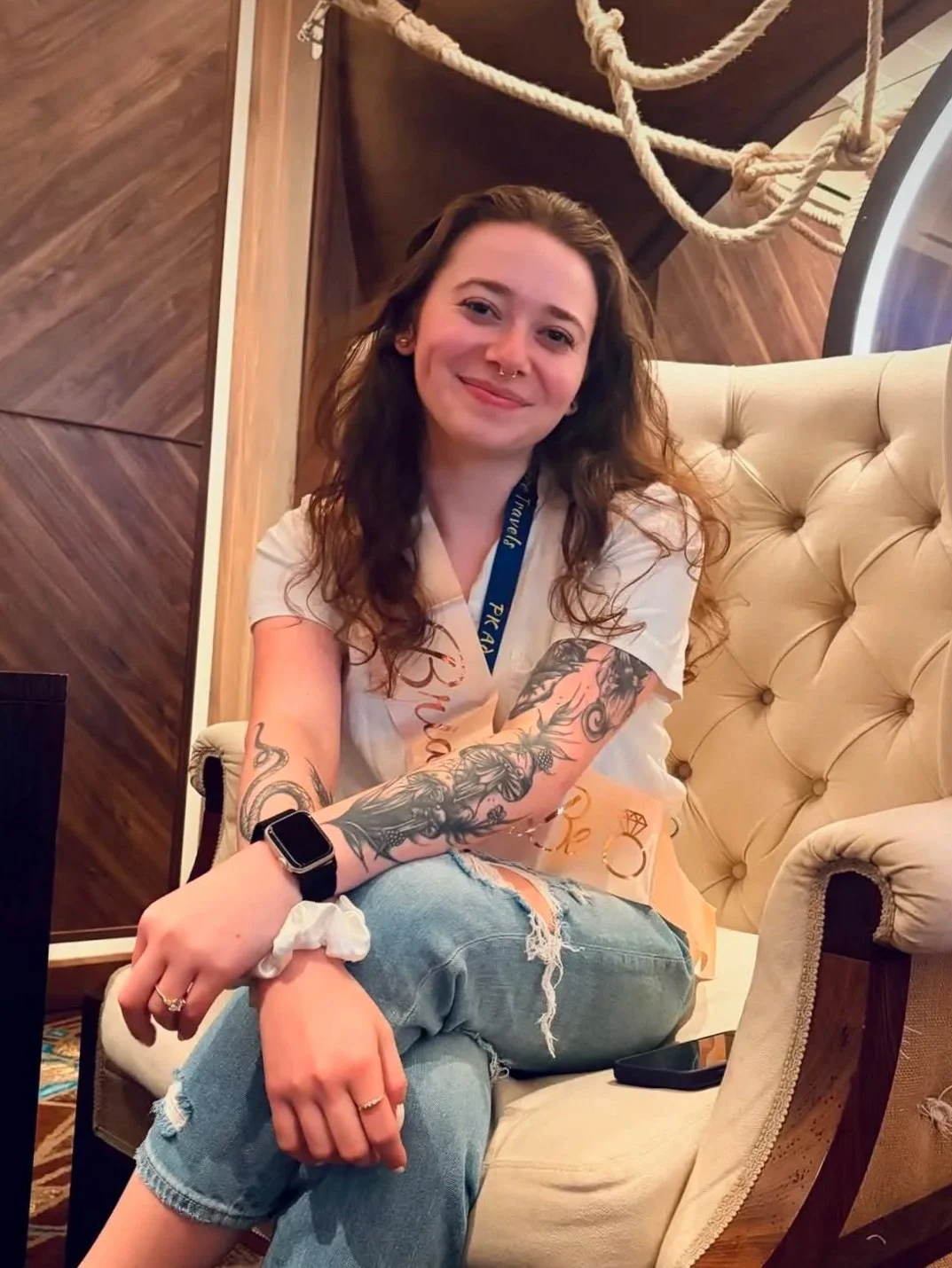 A young woman with curly brown hair and tattoos on her arms sitting on a beige armchair in a cozy room. She's smiling at the camera, wearing a white t-shirt and ripped jeans, with a smartwatch on her left wrist and a ring on her finger.