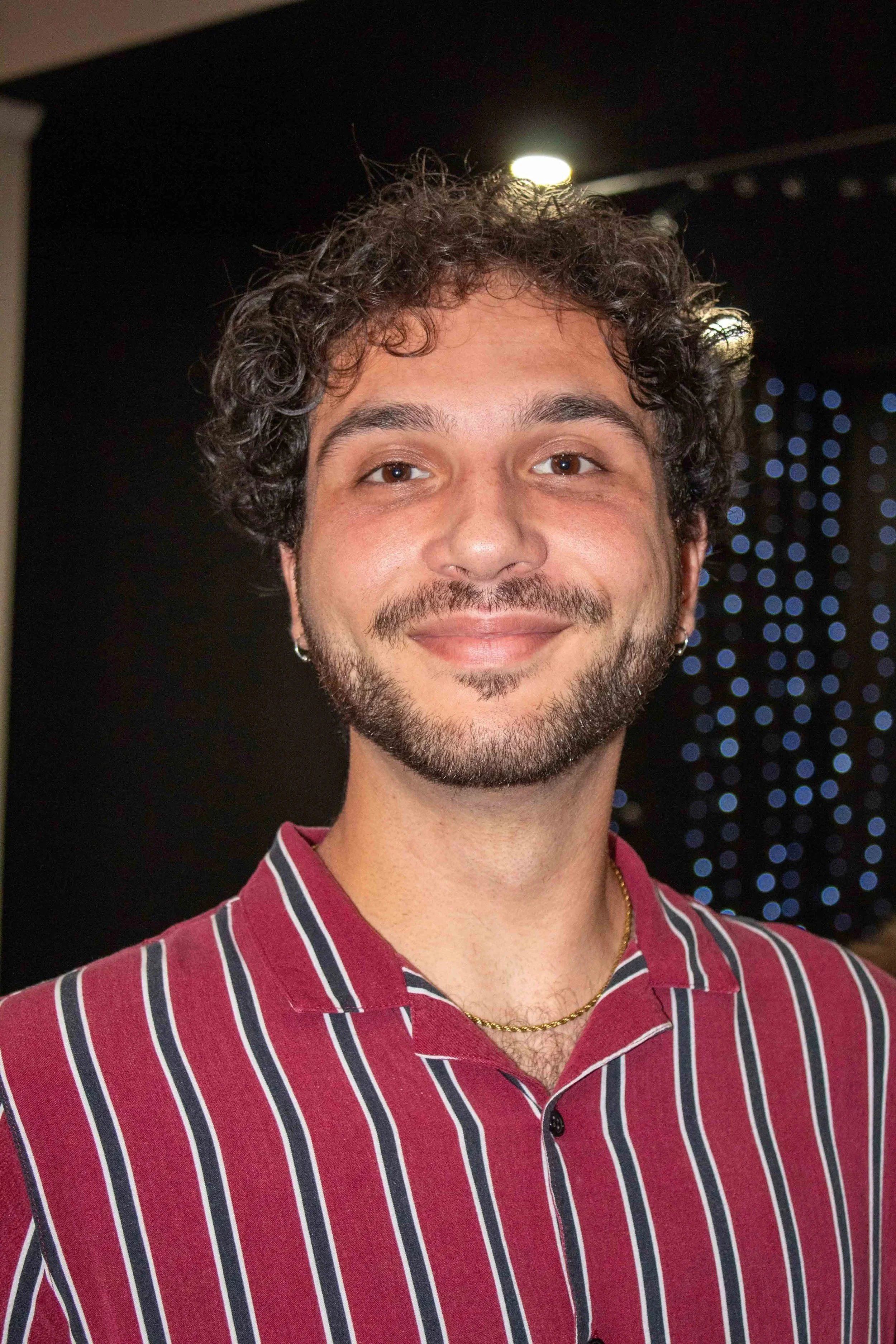 A young man with curly brown hair, a beard, and earrings, smiling at the camera, wearing a maroon striped shirt, with a blurred background of black and blue lights.