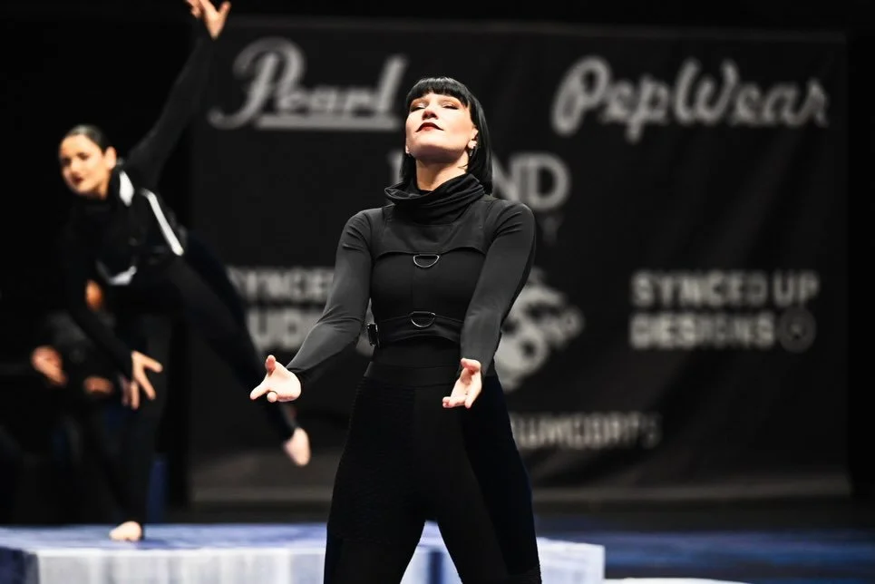 A performer with short black hair and dark makeup, wearing all black, stands on stage with arms extended in a dance pose, with another dancer in the background.